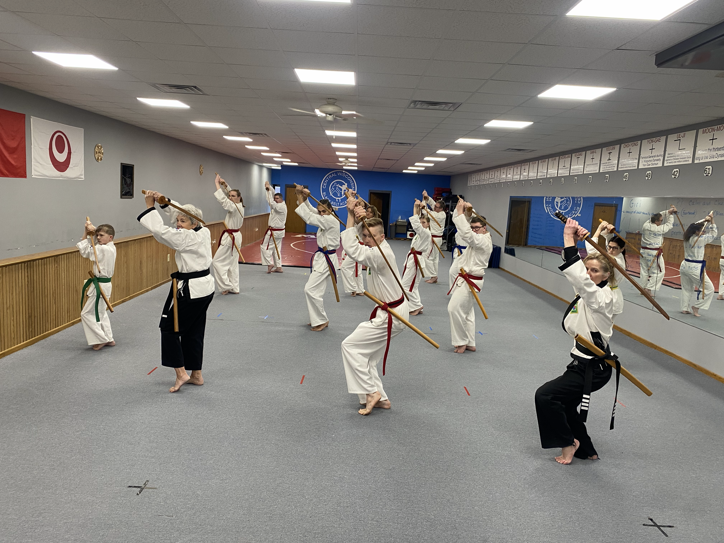 Students practicing martial arts with wooden staffs in a dojo, wearing traditional uniforms and belts, in front of a mirror and a blue wall with martial arts symbols.
