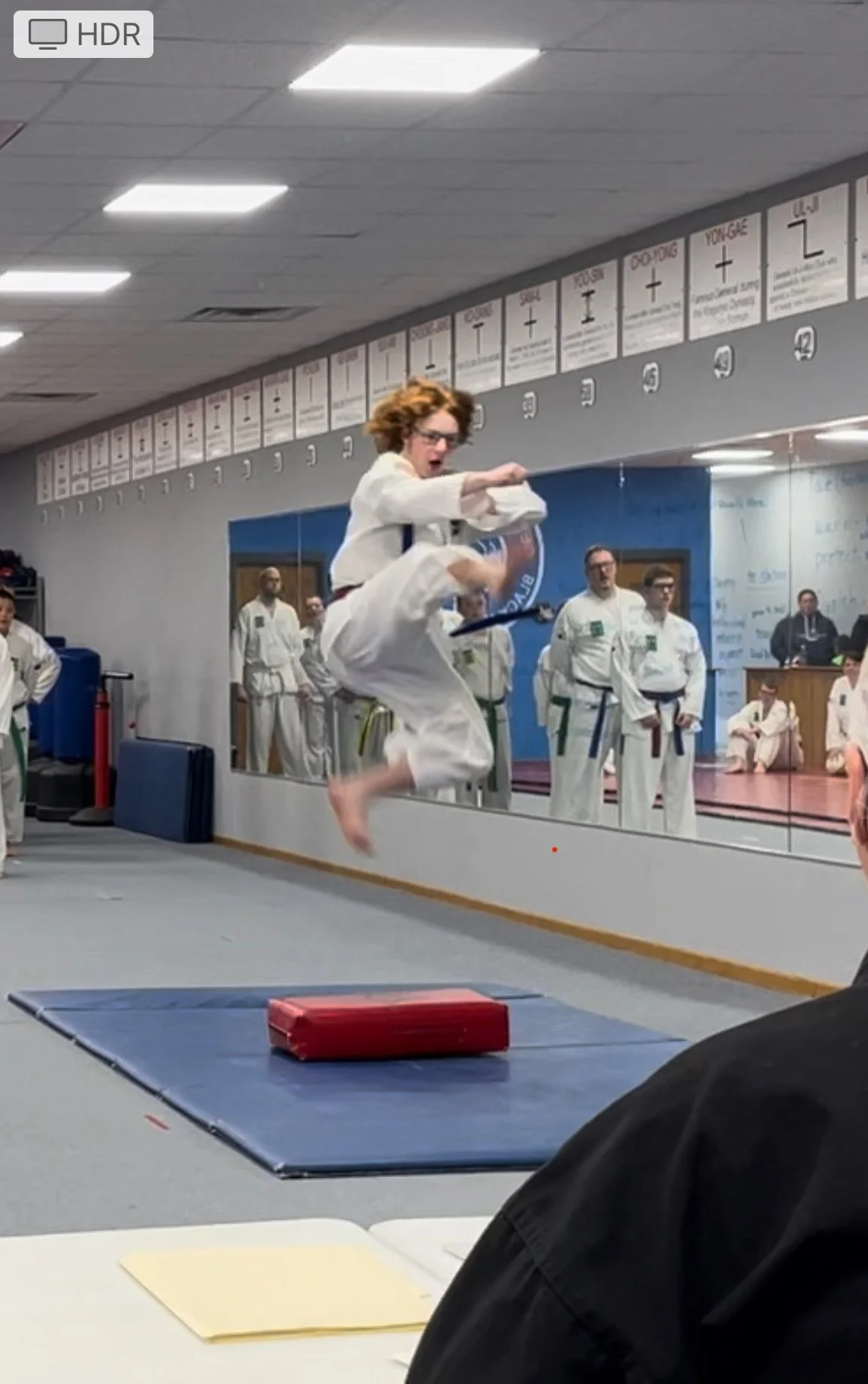 A martial arts student in a white gi is mid-air performing a kick during training or demonstration at a dojo or martial arts school, with onlookers and instructors observing in the background.