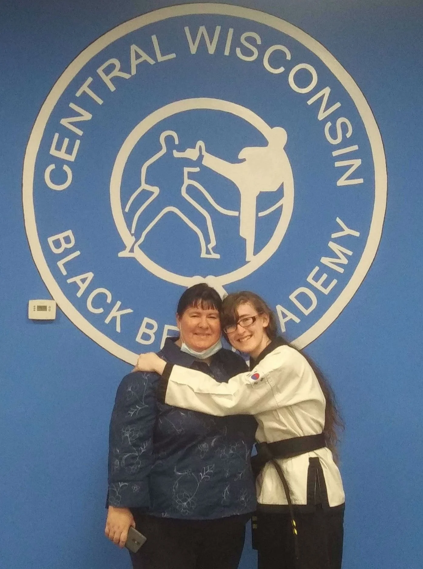 Two women hugging in front of a large blue wall with a martial arts logo that says 'Central Wisconsin Black Belt Academy,' featuring two martial artists in a fighting stance.