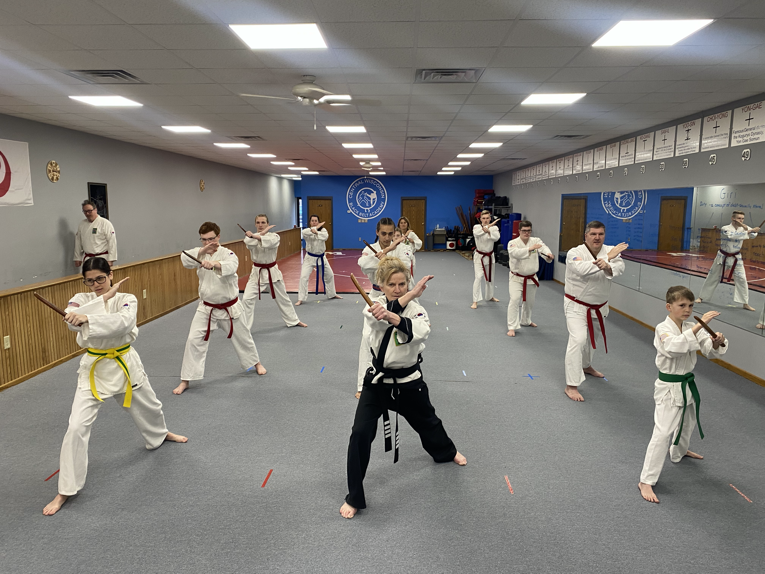 Martial arts class with students in white uniforms practicing with wooden swords in a gymnasium, led by an instructor in black uniform.