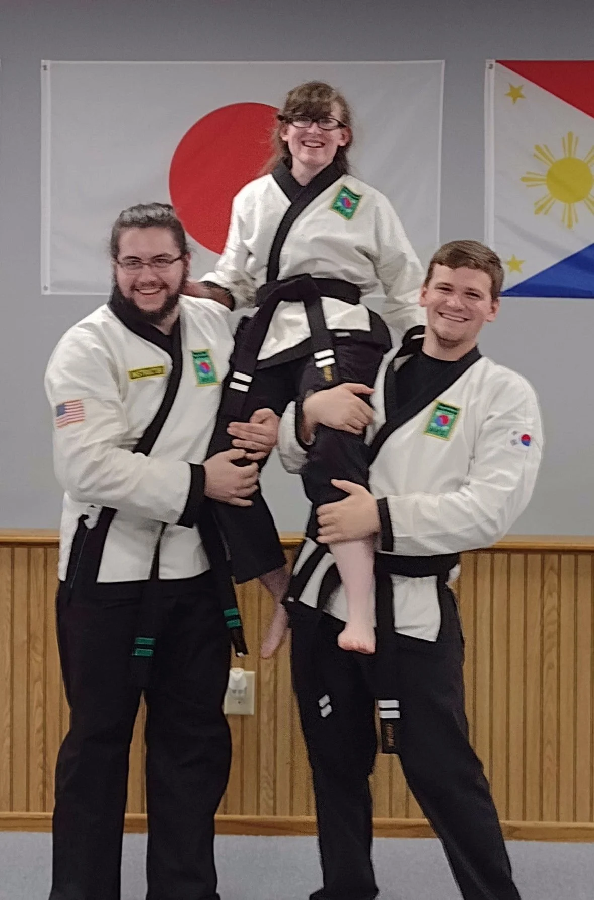 Three martial artists in uniforms with patches, two men , holding a woman with a black belt, all smiling, in front of Japanese and Philippine flags.