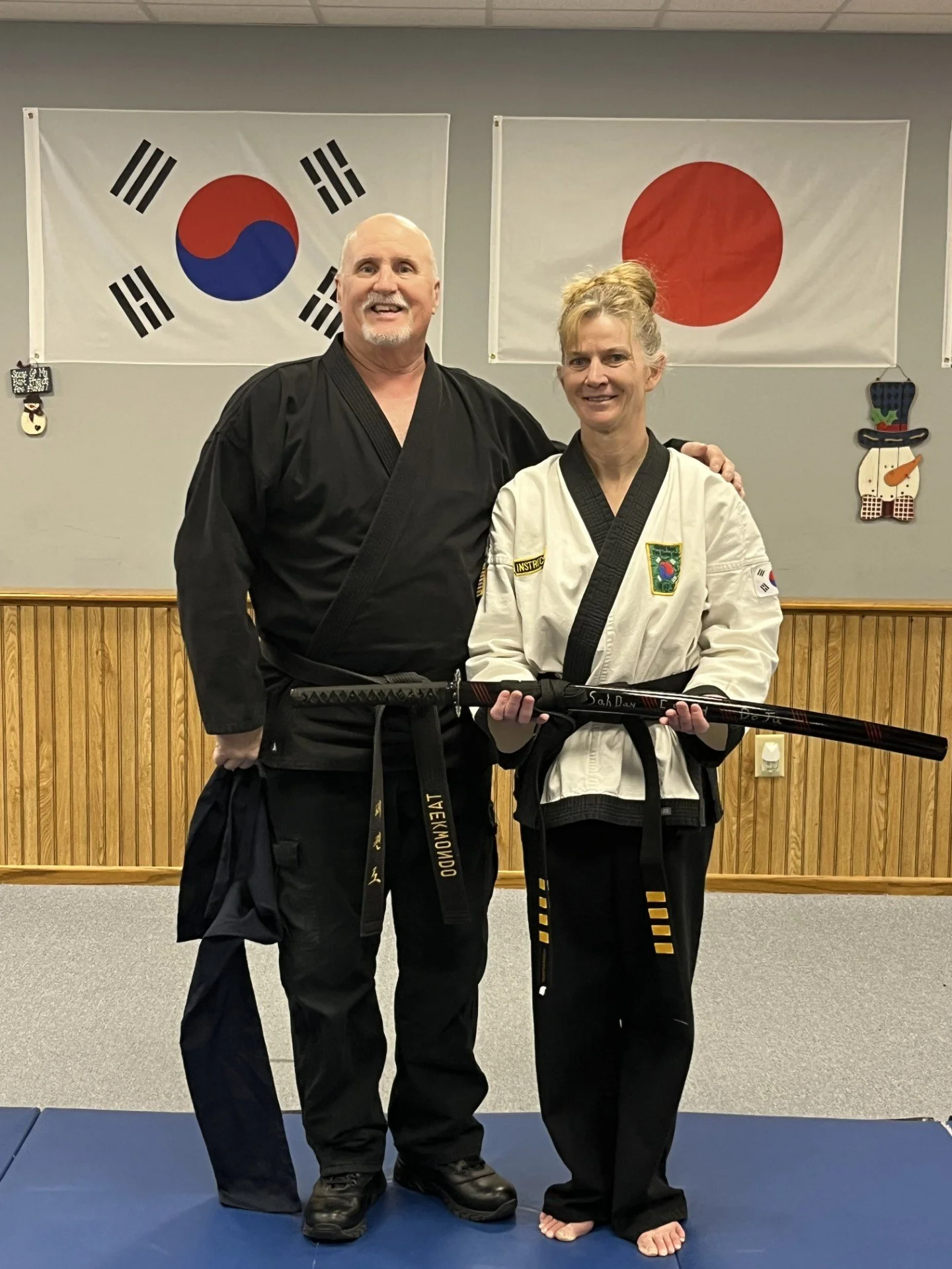 A man and woman in martial arts uniforms standing in a dojo, holding a martial arts sword together. Two flags, South Korea and Japan, hang on the wall behind them.