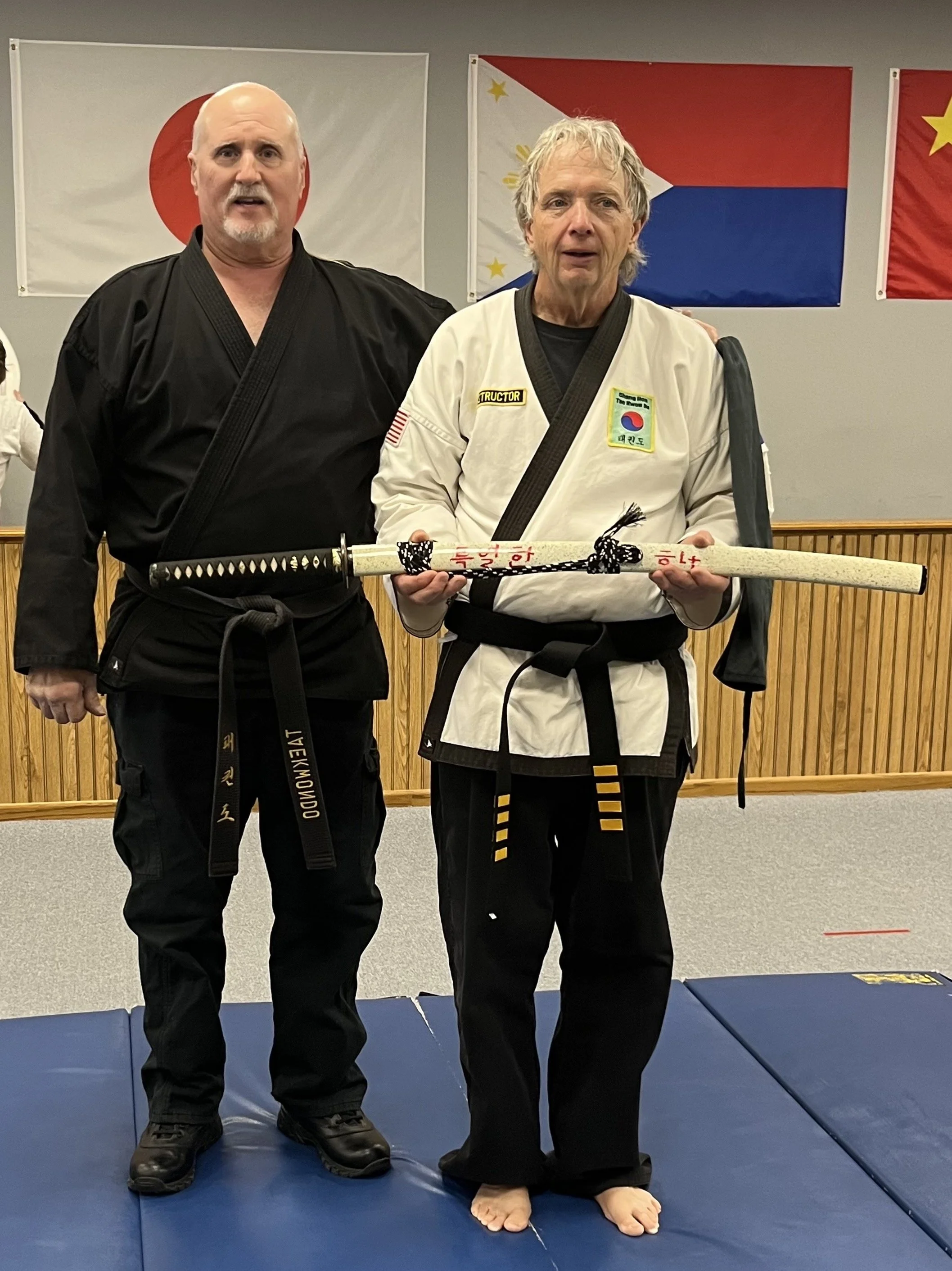 Two martial artists in uniforms, standing on a blue mat indoors, with flags hanging on the wall behind them. The person on the right holds a gifted sword.