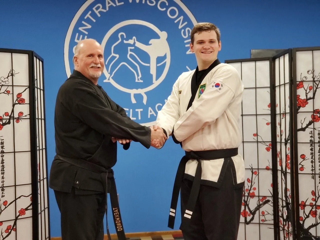 Two men in martial arts uniforms shaking hands inside a martial arts dojo. The background features the logo of the United States Taekwondo Union and folding screens with cherry blossom designs.