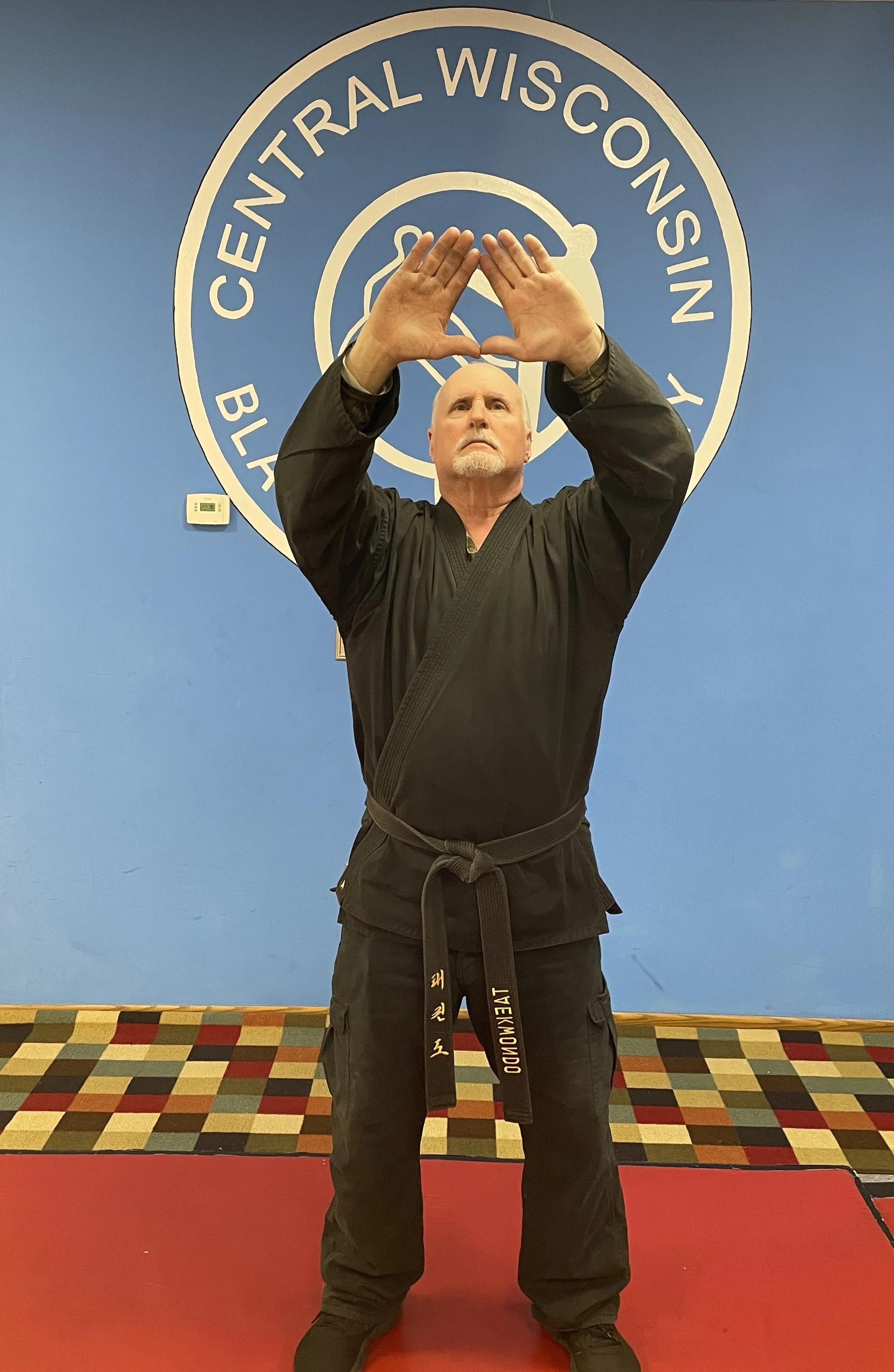 A man in a black martial arts uniform standing on a red mat, forming a triangle with his hands near his forehead. Behind him is a blue wall with the logo of Central Wisconsin Black Belt Academy.
