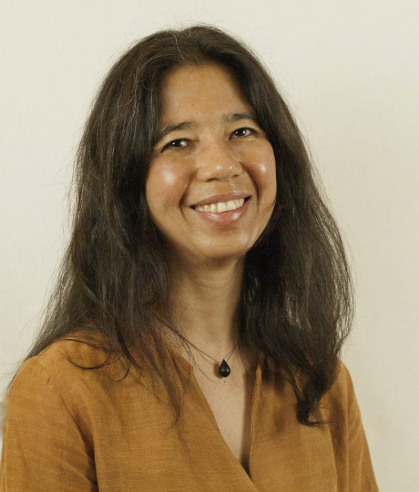 Portrait of a woman with long dark hair wearing a mustard-colored top and a black pendant necklace, smiling at the camera.