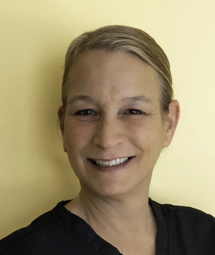 Close-up of a woman with short blonde hair smiling, wearing a black top, against a light yellow background.