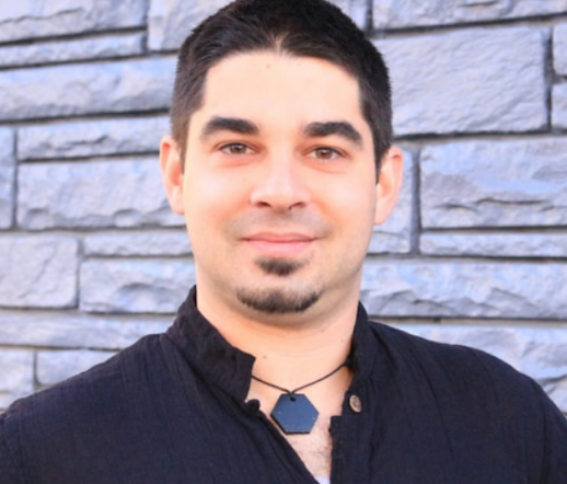 A young man with short dark hair and a goatee smiling in front of a gray stone wall, wearing a black shirt and a black necklace.