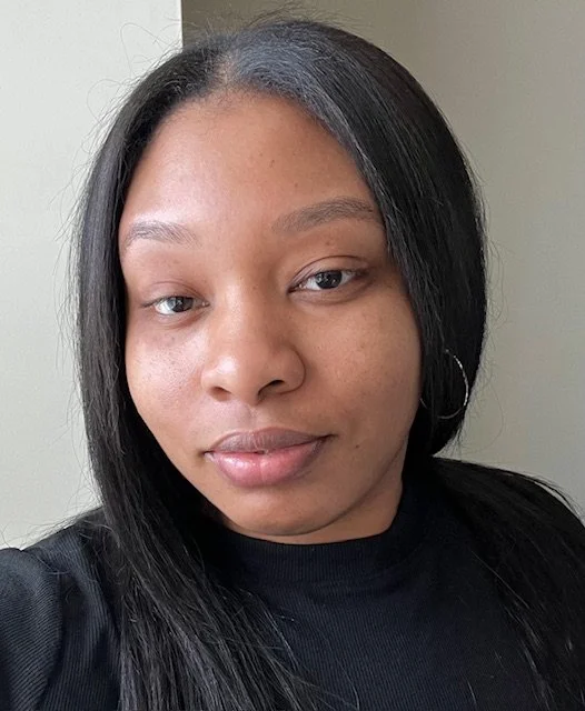 Close-up of a woman with straight black hair, wearing hoop earrings and a black top, looking at the camera with a neutral expression.