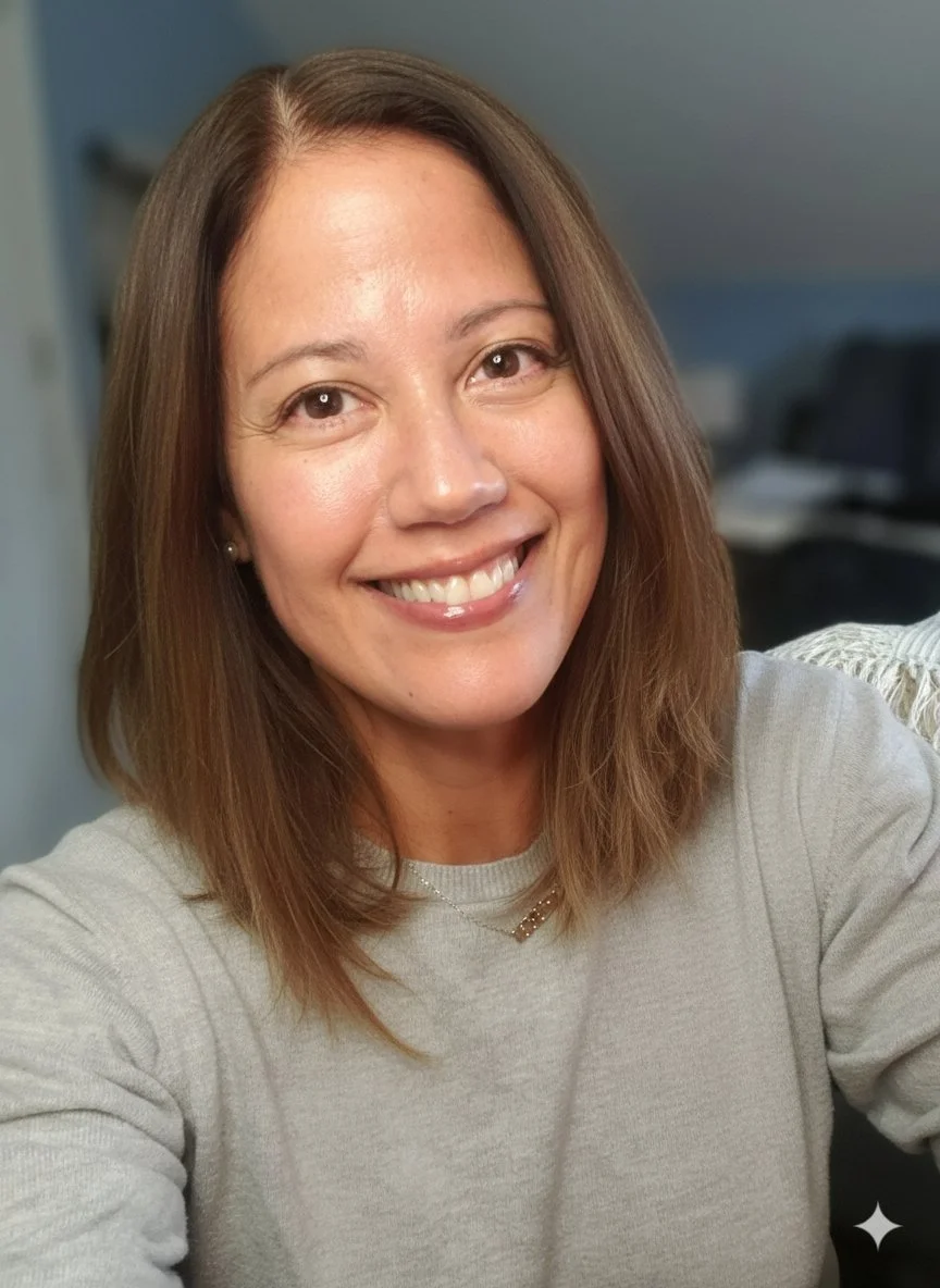 A woman with shoulder-length brown hair smiling at the camera, wearing a light-colored sweater and small earrings, in a room with a blue wall and blurred furniture in the background.