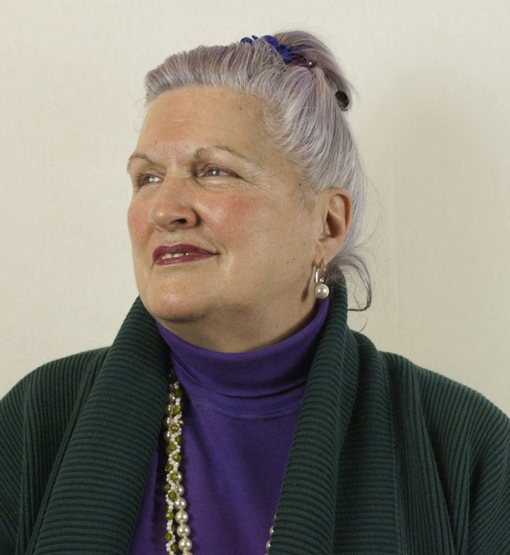 A senior woman with gray hair in a ponytail, wearing pearl earrings, a purple turtleneck, a green jacket, and a pearl necklace, looking slightly to the side.