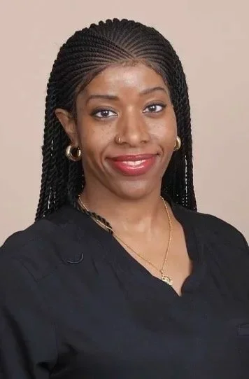A woman with braided hair, wearing gold hoop earrings, a nose ring, a gold necklace, and a black top, smiling at the camera.