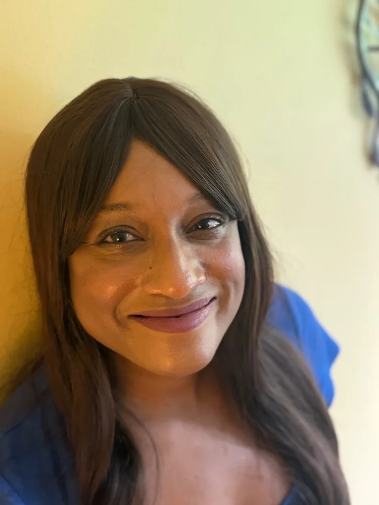 A woman with long brown hair and a blue top smiling and leaning against a yellow wall.