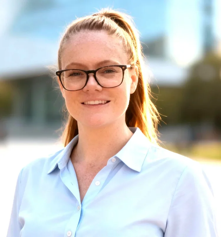 Close-up of a young woman with red hair tied in a high ponytail, wearing glasses and a light blue collared shirt, smiling outdoors with a blurred cityscape background.