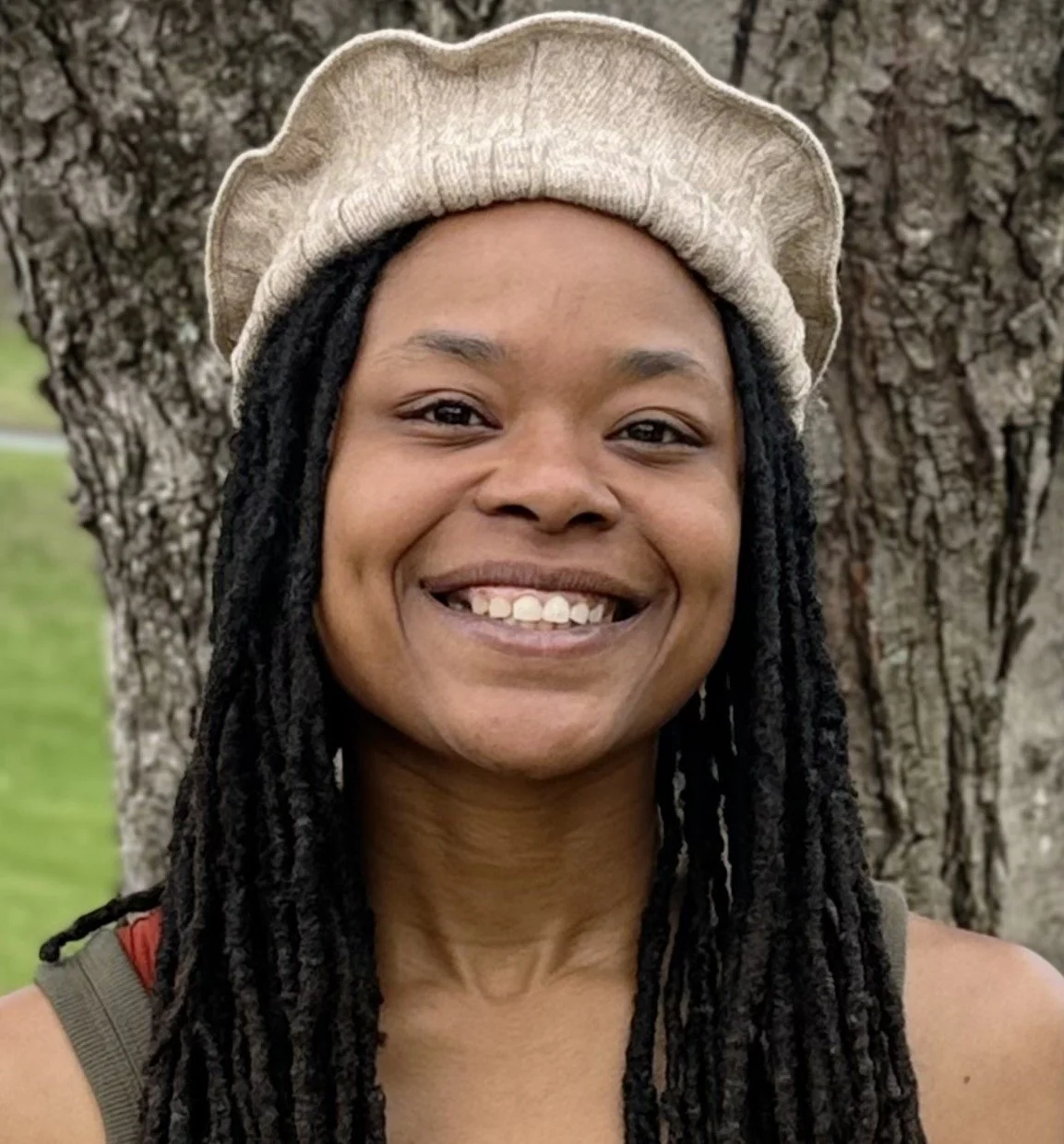 Smiling woman with long black dreadlocks wearing a beige beret standing outdoors in front of a tree.