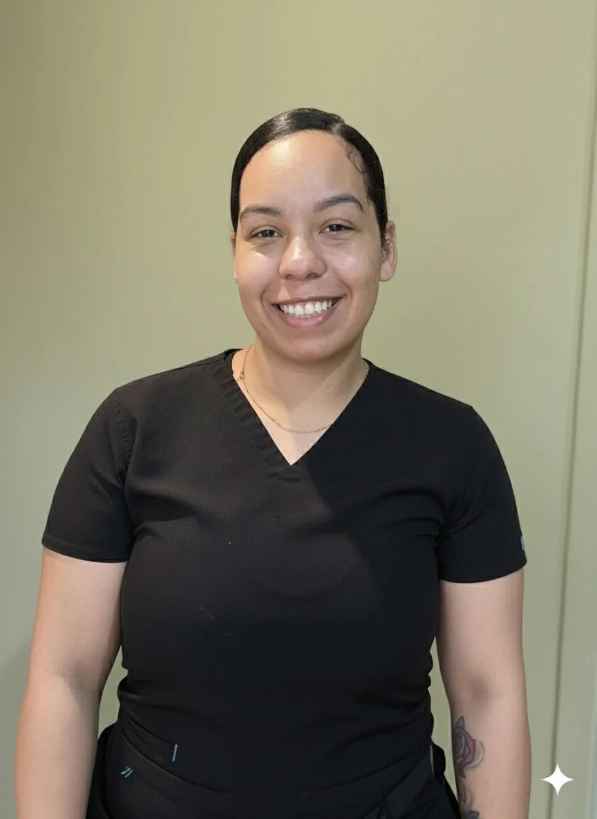 A woman with dark hair pulled back, wearing a black scrub top and smiling, standing in front of a plain, light-colored wall.