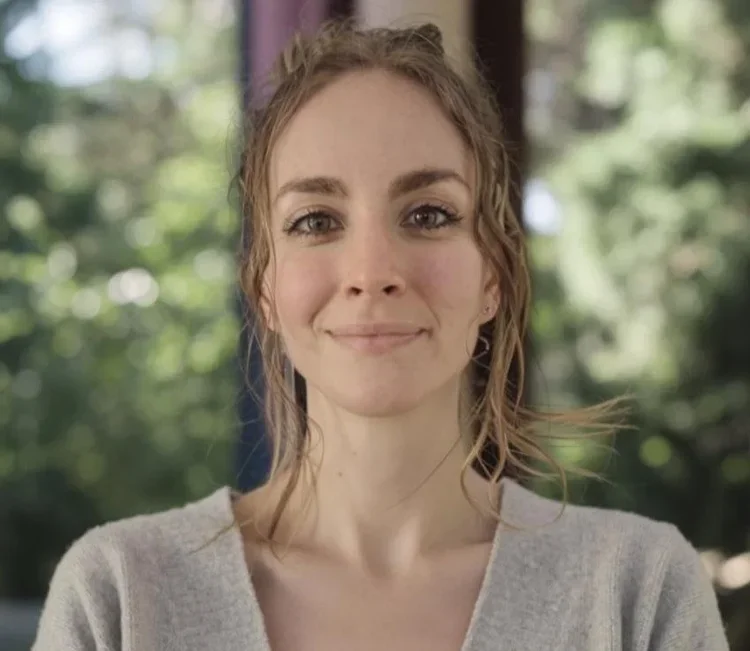 A woman with shoulder-length wavy brown hair smiling outdoors with a blurred background of trees.