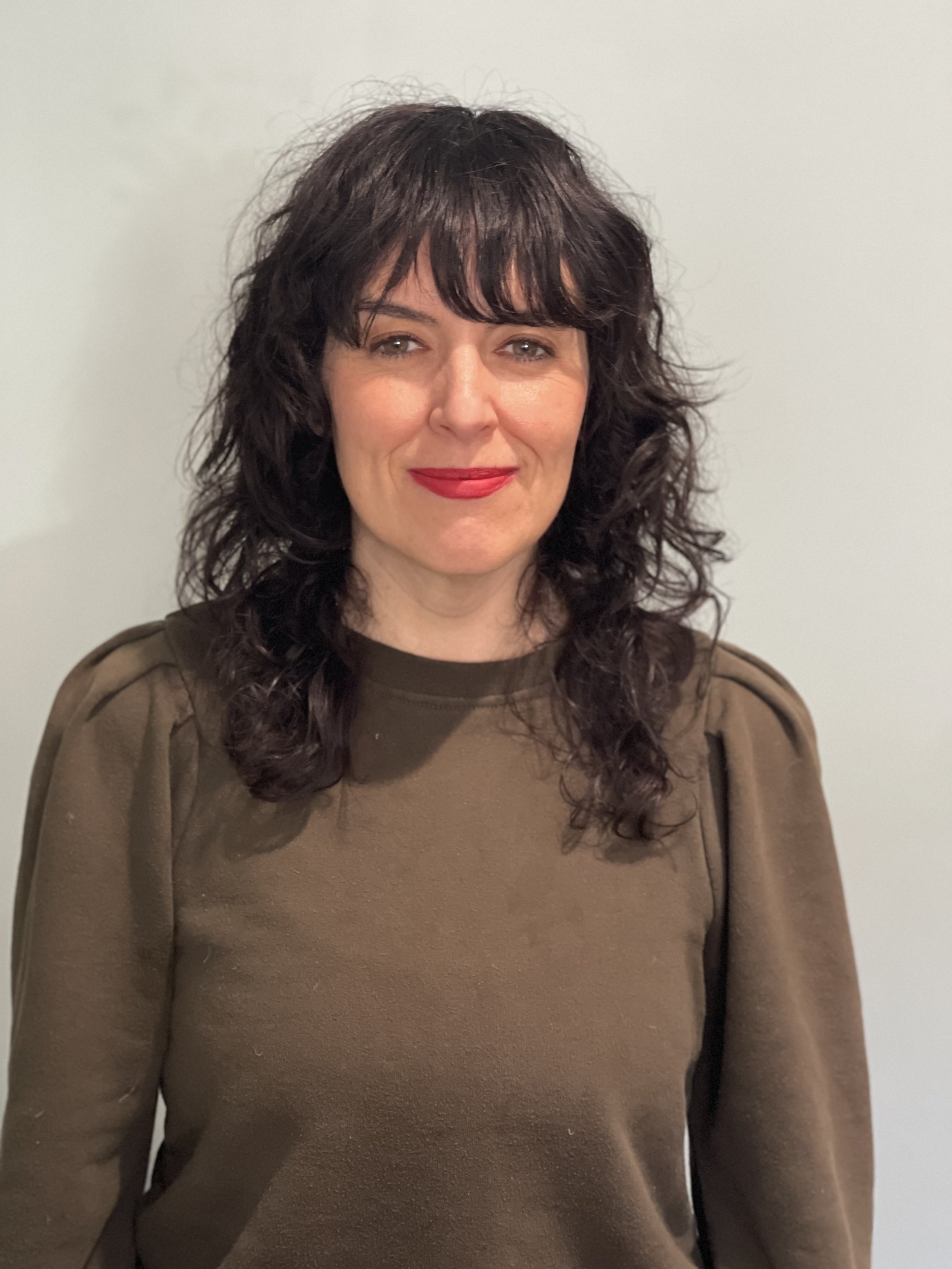 A woman with curly dark hair and red lipstick, wearing a brown top, standing against a plain light-colored wall.