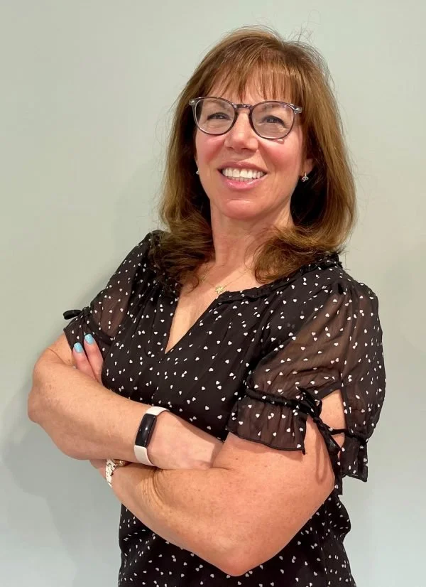 A smiling woman with shoulder-length auburn hair, wearing glasses and a black blouse with white polka dots, standing with her arms crossed against a plain light-colored background.