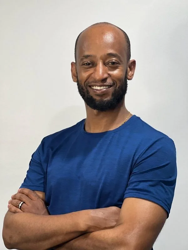 A smiling man with a beard and shaved head, wearing a blue t-shirt, standing with arms crossed against a plain white wall.