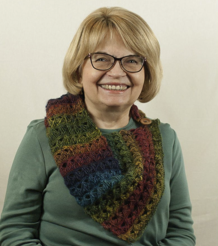 A smiling woman with blonde hair and glasses wearing a green top and a multicolored scarf.
