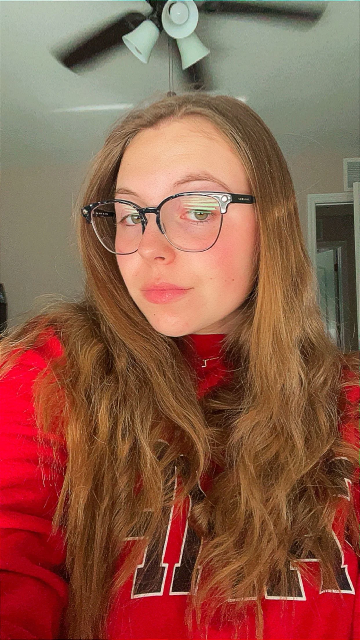 A young woman with long, wavy brown hair wearing glasses and a red sweatshirt, indoors, with a ceiling fan in the background.