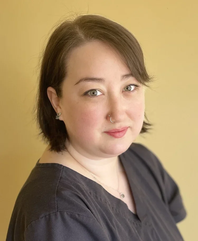 A woman with short brown hair, grey eyes, and light skin, wearing a black shirt and jewelry, standing in front of a yellow background.