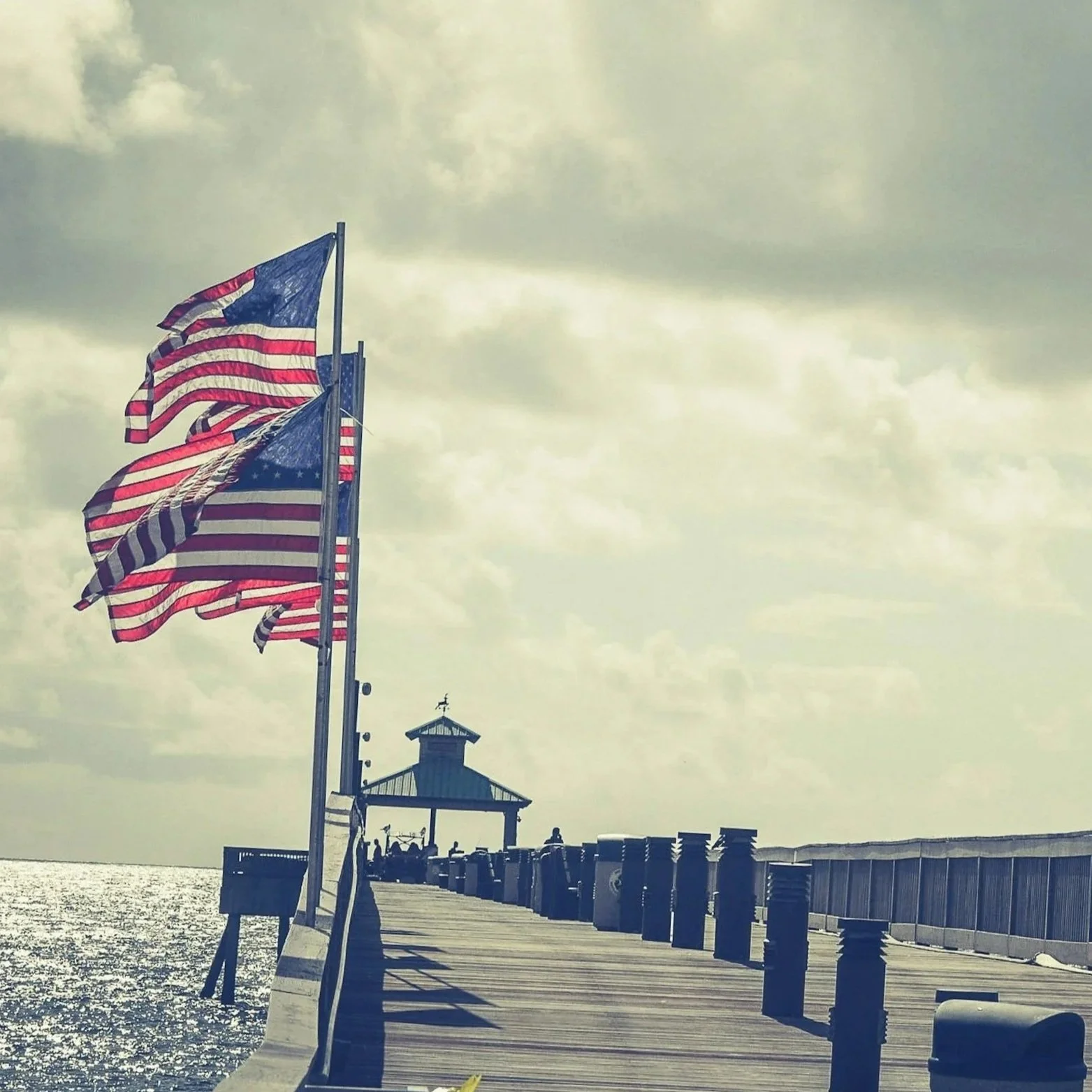 American flags waving on a pier with a gazebo at the end, overlooking the water on a cloudy day.