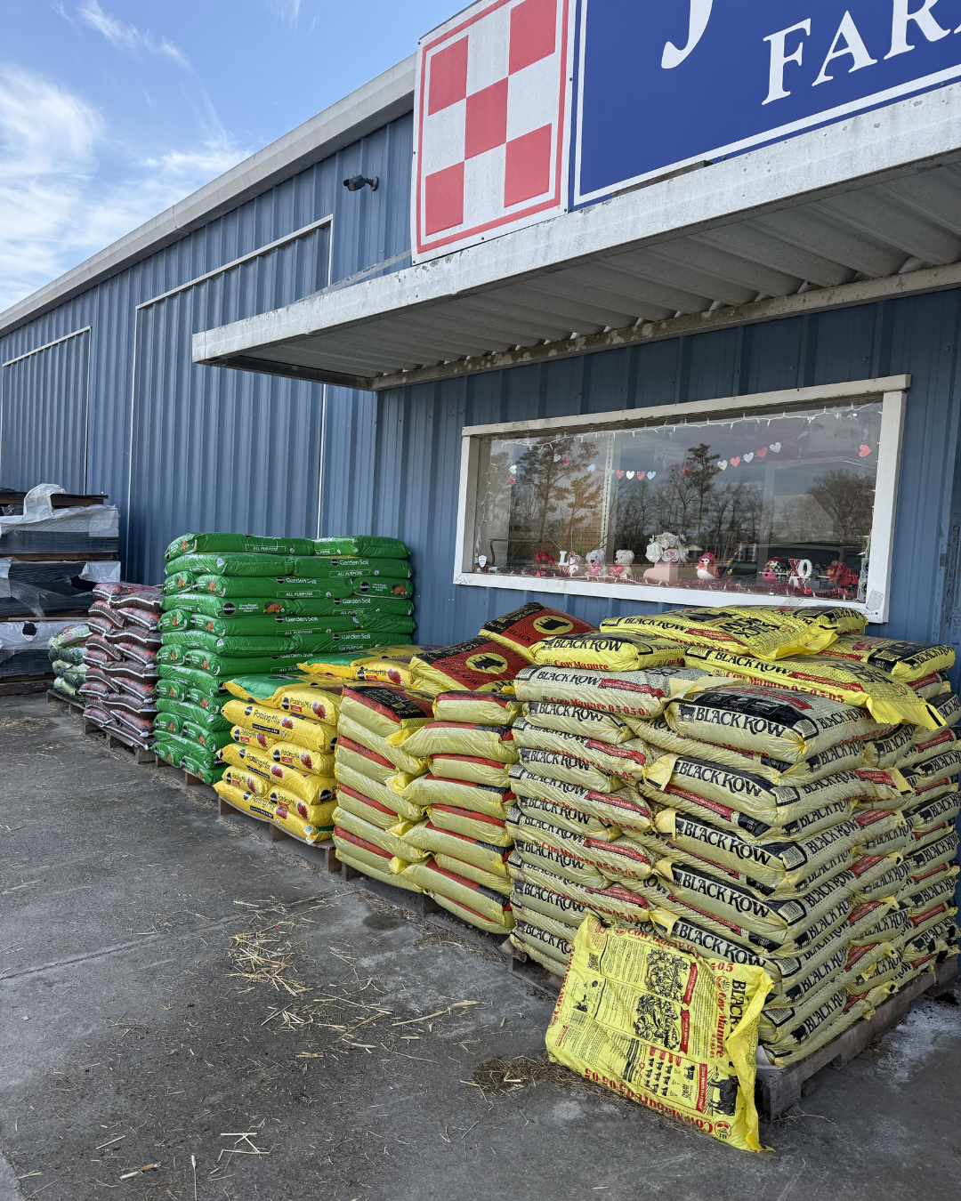 Piles of garden soil and fertilizer bags outside a store with blue metal siding, a window decorated with small heart-shaped cutouts, and a sign with a red and white checkered pattern.