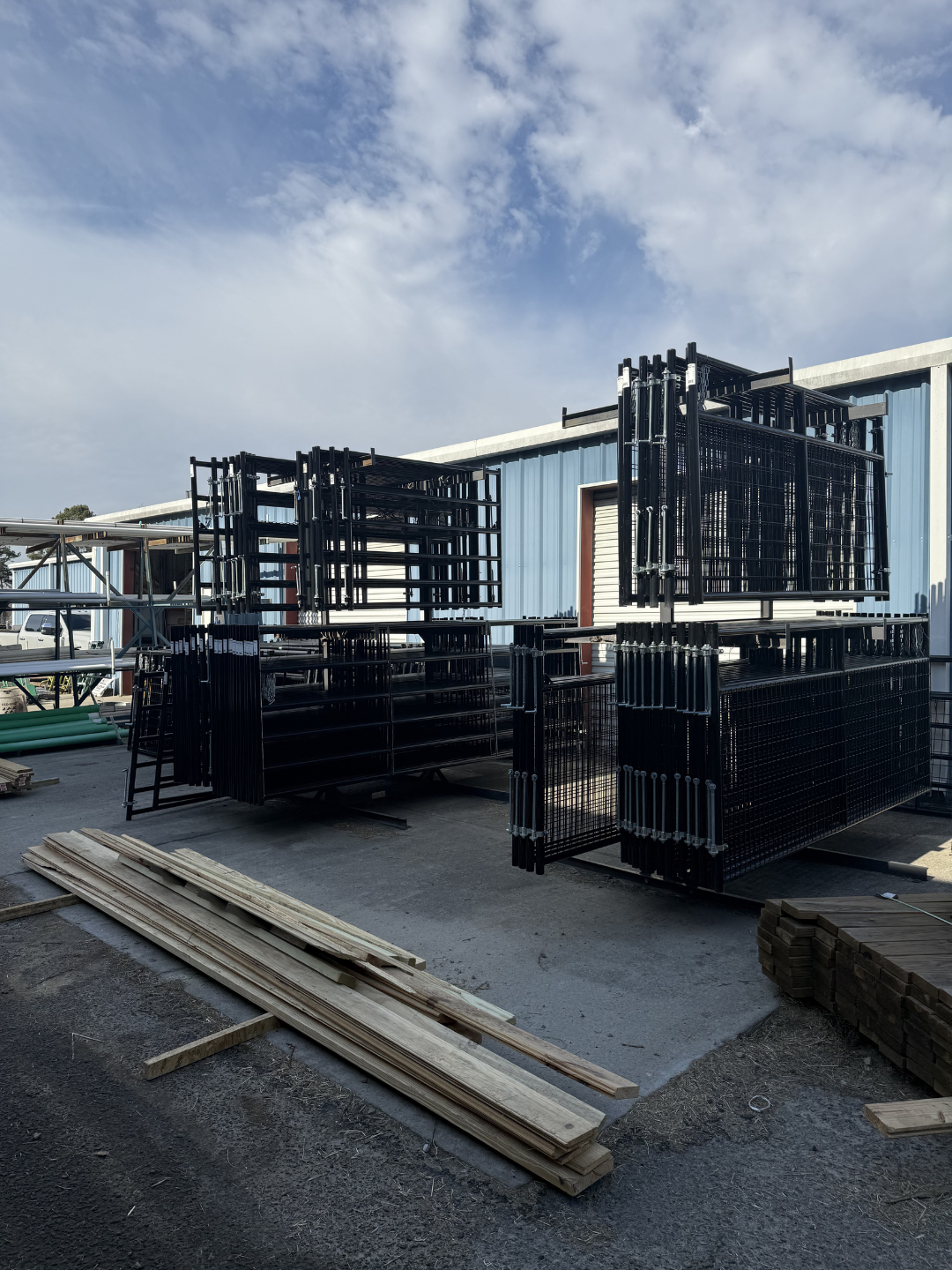 Stacks of black metal fencing panels and wooden planks outdoors near a blue industrial building under a partly cloudy sky.