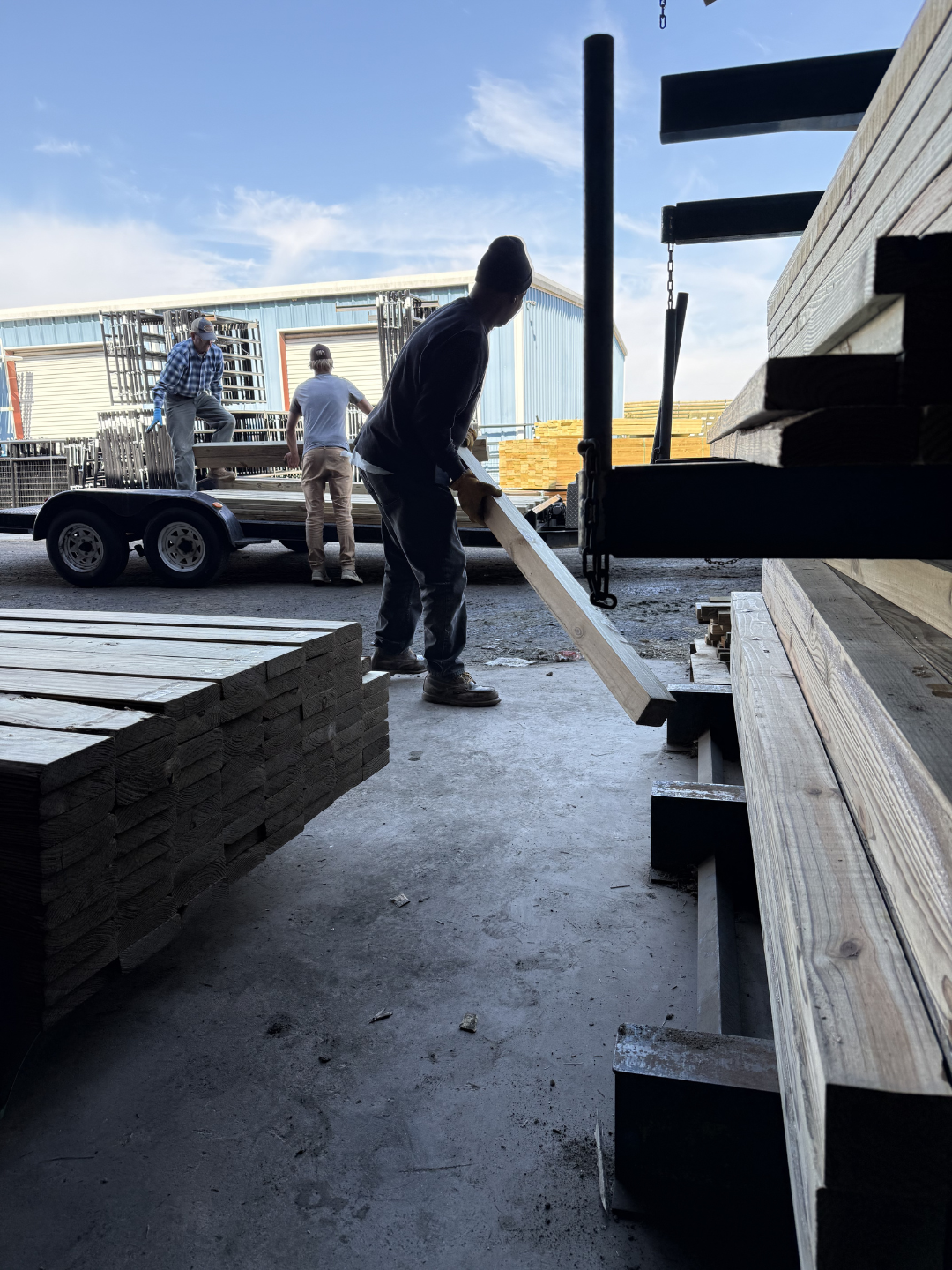 Workers loading lumber onto a trailer outside a warehouse on a sunny day.