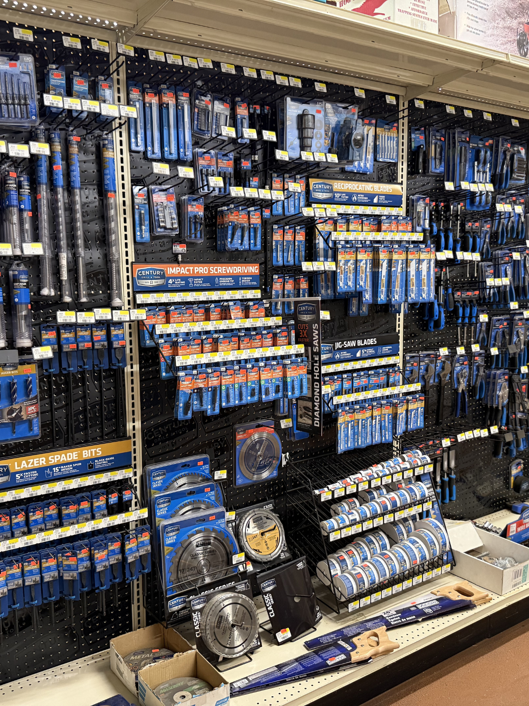 A store shelf displays various hand tools and accessories, including saw blades, screwdrivers, bits, and blades organized on black pegboards with yellow price tags.