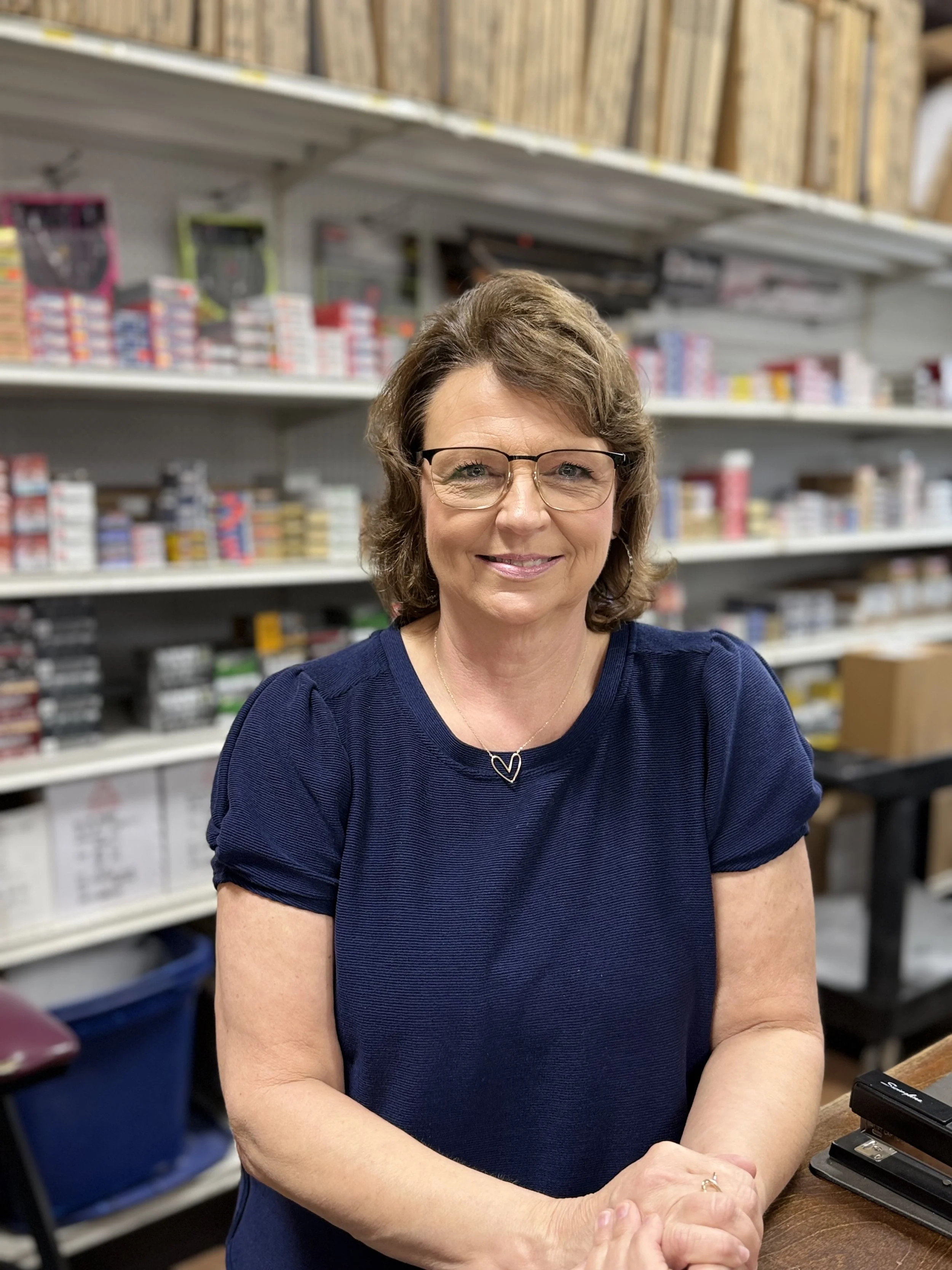 A woman with glasses and curly brown hair smiling, wearing a navy blue shirt, sitting at a counter in a store with shelves of small boxes and products in the background.