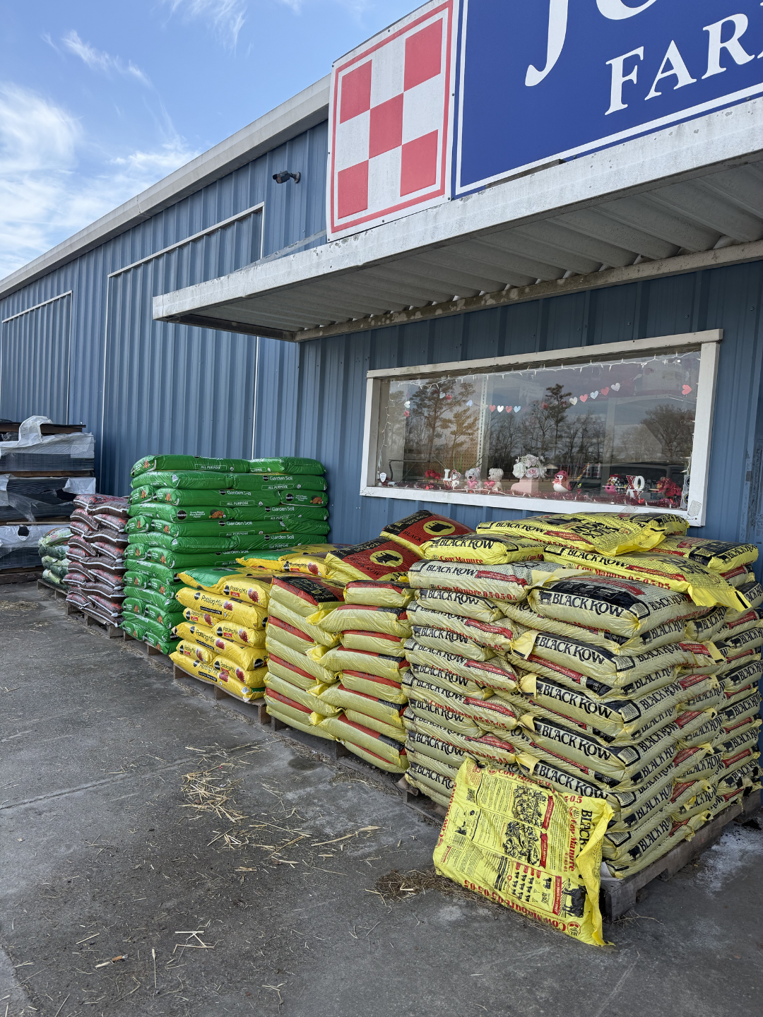 Stacked bags of gardening soil and fertilizer outside a store with a blue exterior and a window, under a partly cloudy sky.