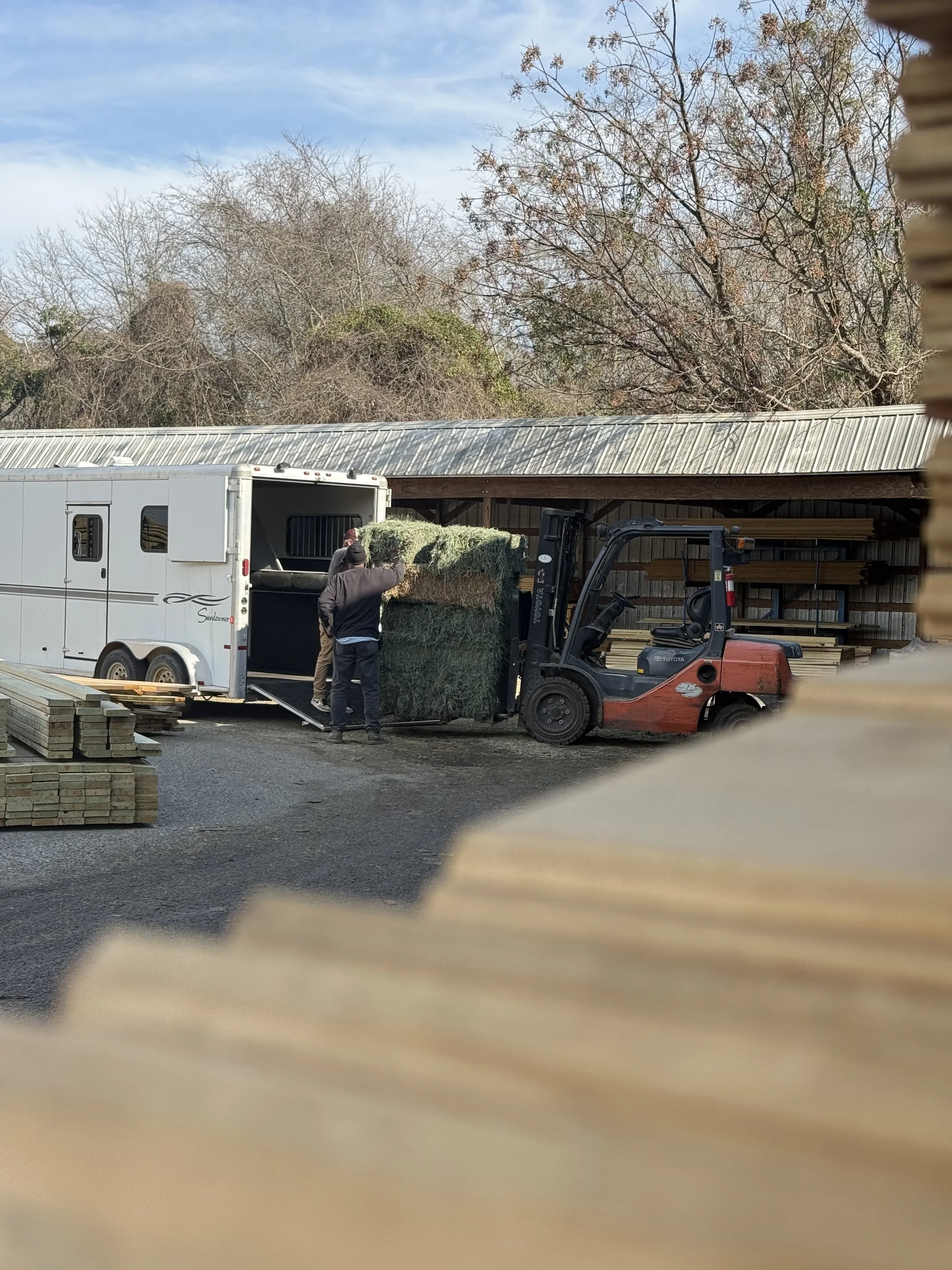 Two people loading hay bales from a trailer into a forklift outside a wooden structure with trees in the background.