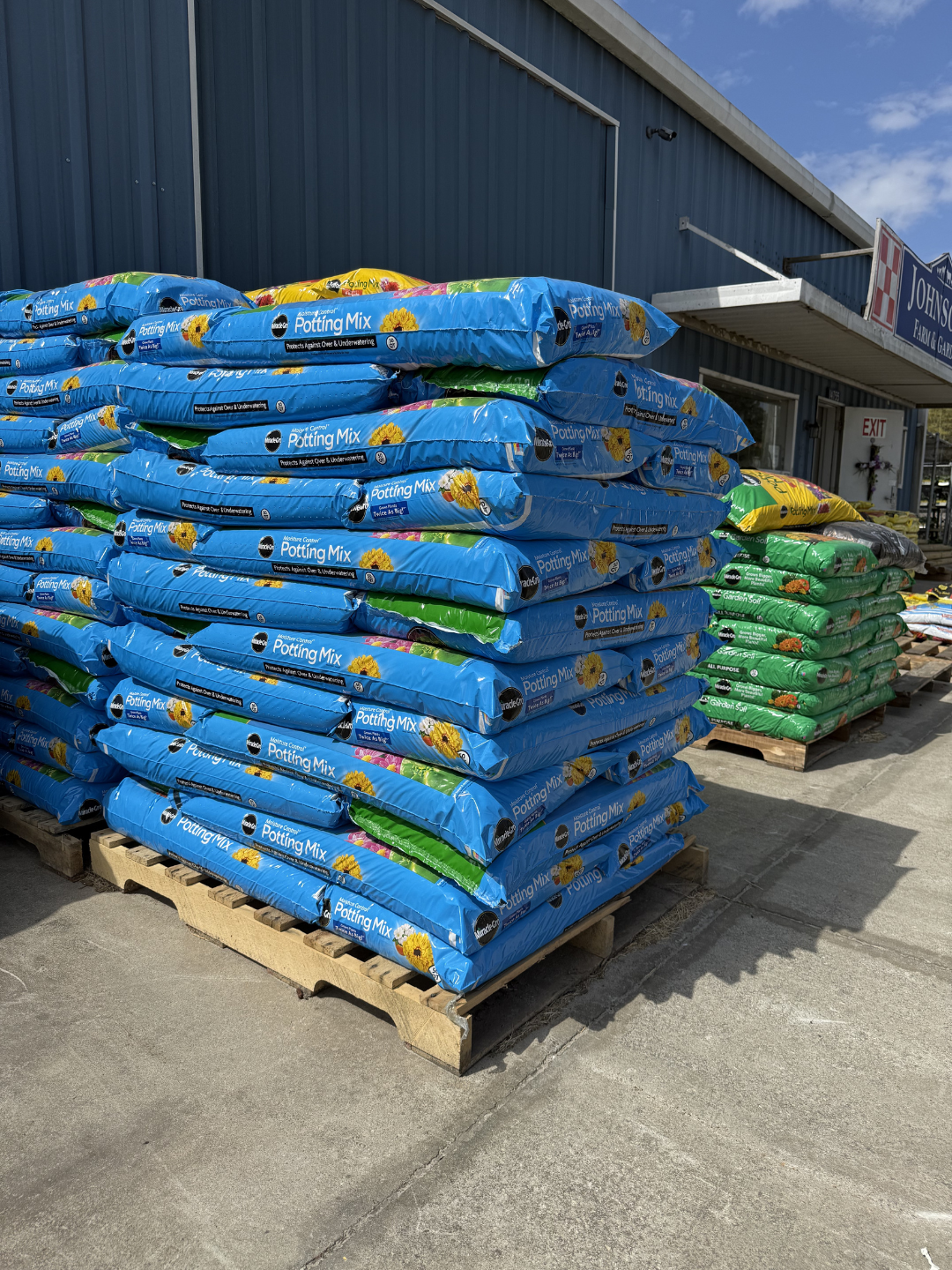 Piles of bags of potting mix on wooden pallets outside store, blue and green bags, store building and sign in background, clear sky.