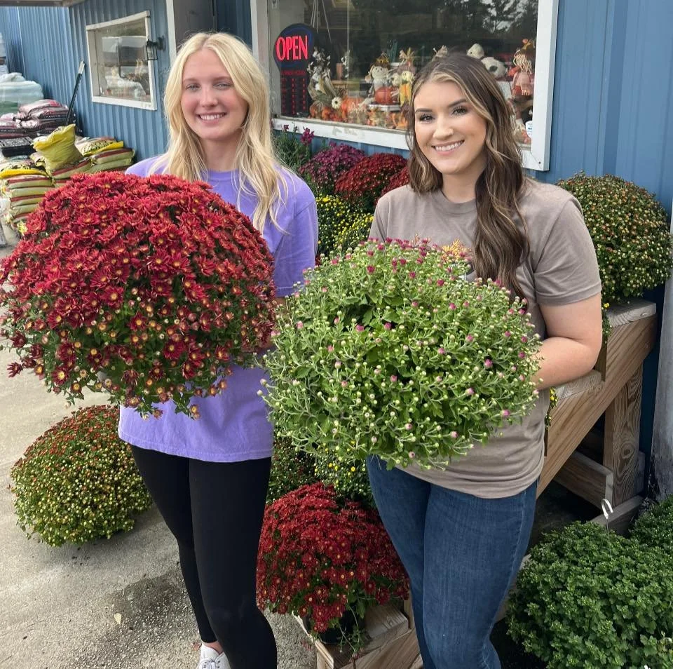Two women smiling and holding potted chrysanthemums in front of a flower shop. The shop has various colorful potted plants and flowers, with an 'Open' sign in the window.