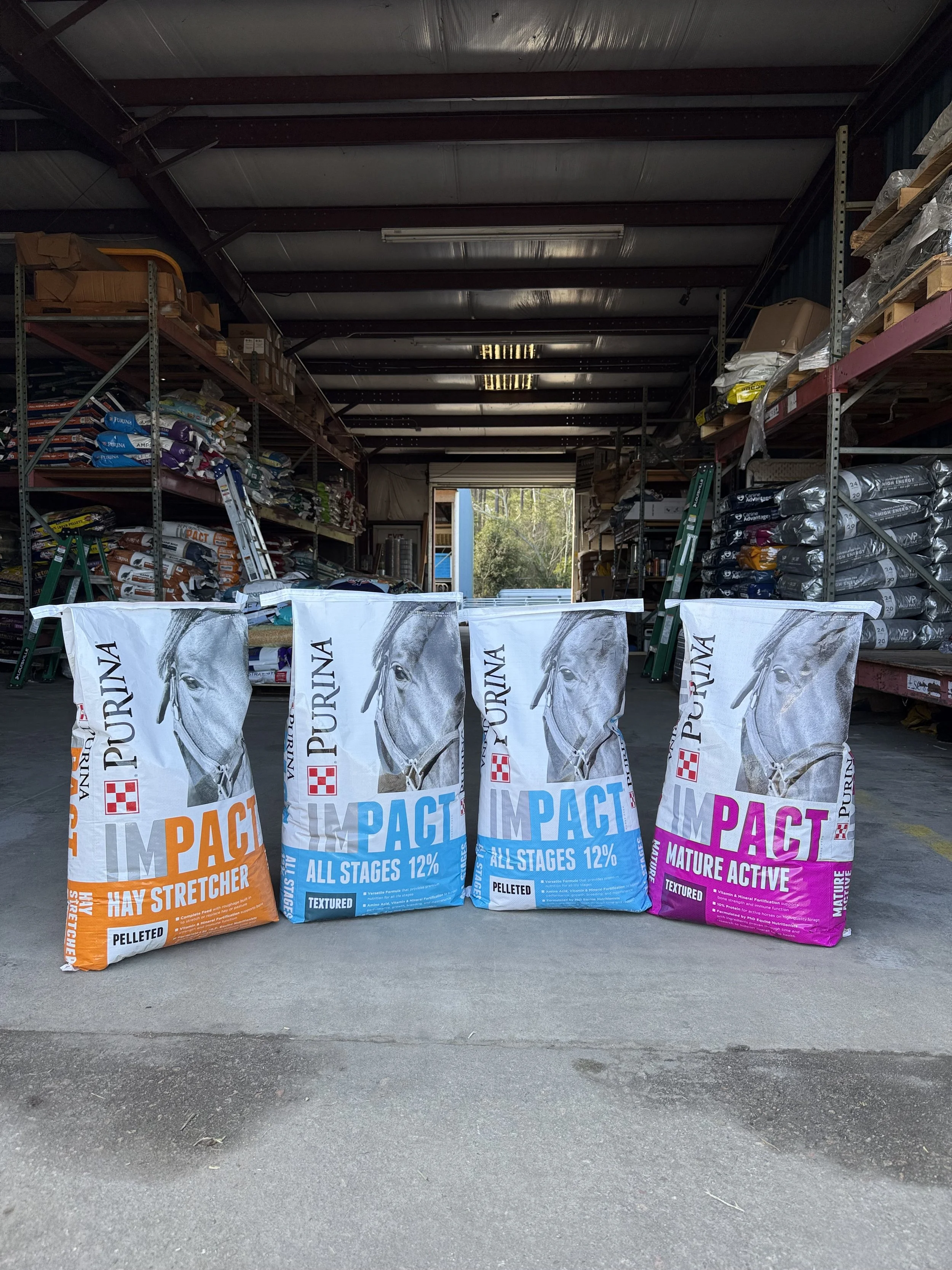 Four large bags of horse feed labeled Purina Impact, with different varieties, in a warehouse aisle.