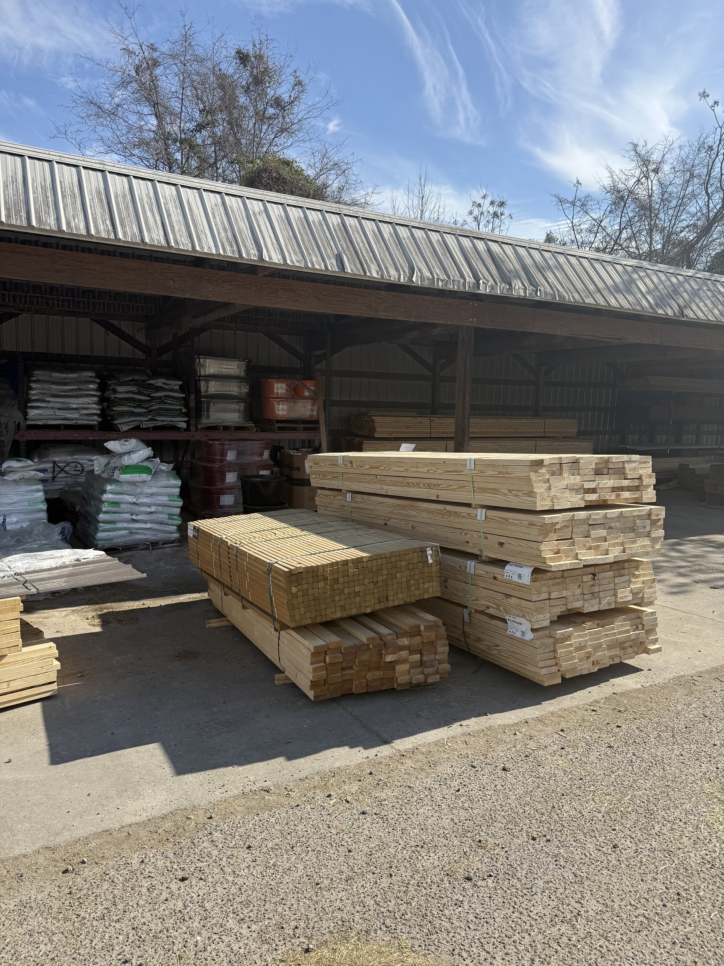 Stacks of lumber and wood planks stored outdoors under a metal roof, with bags of other materials and trees visible in the background.