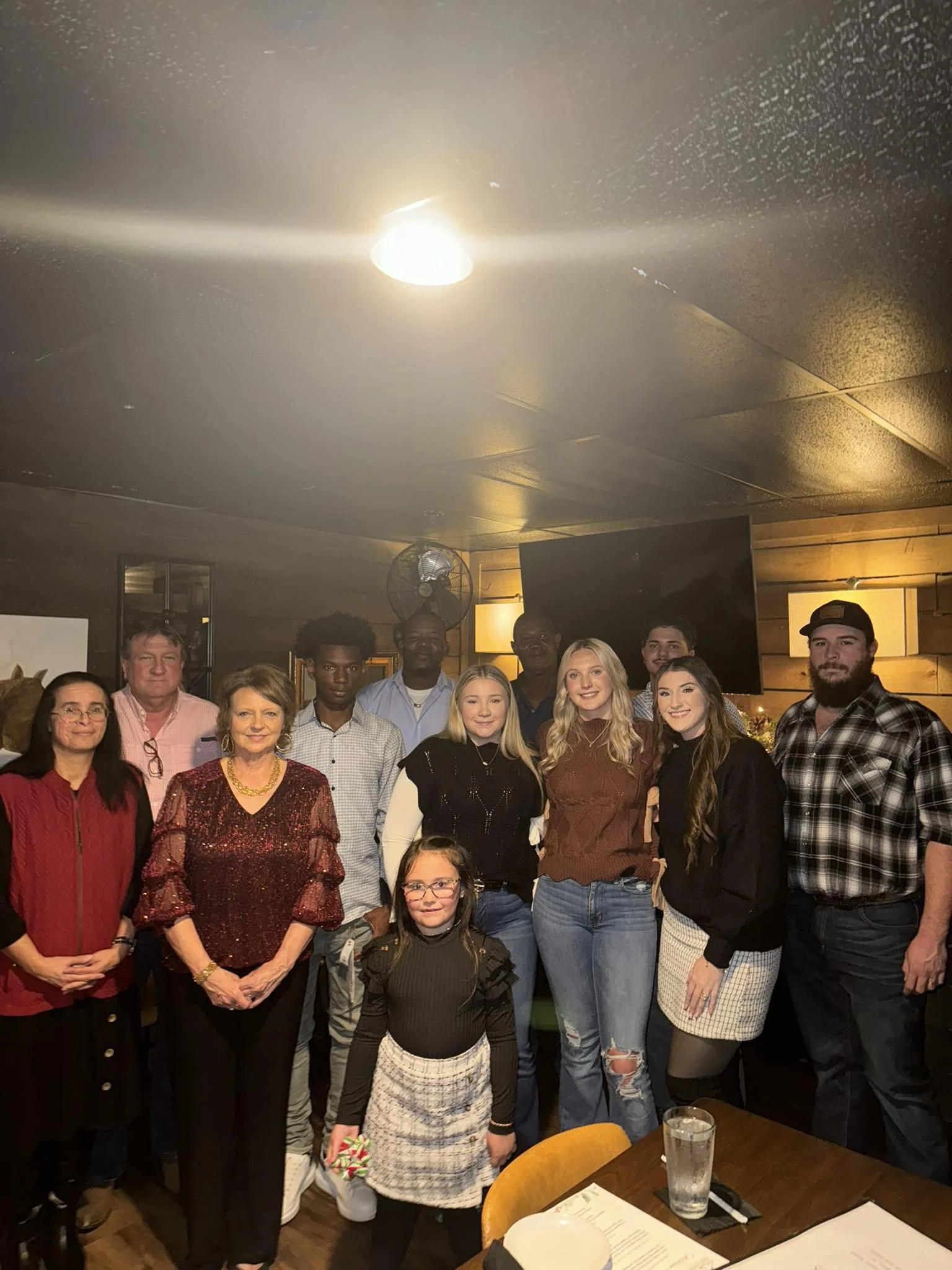 A group of ten people, including children and adults, gathered together for a photo in a restaurant or banquet room. They are standing in front of a wooden wall with a TV and a fan in the background, smiling at the camera.