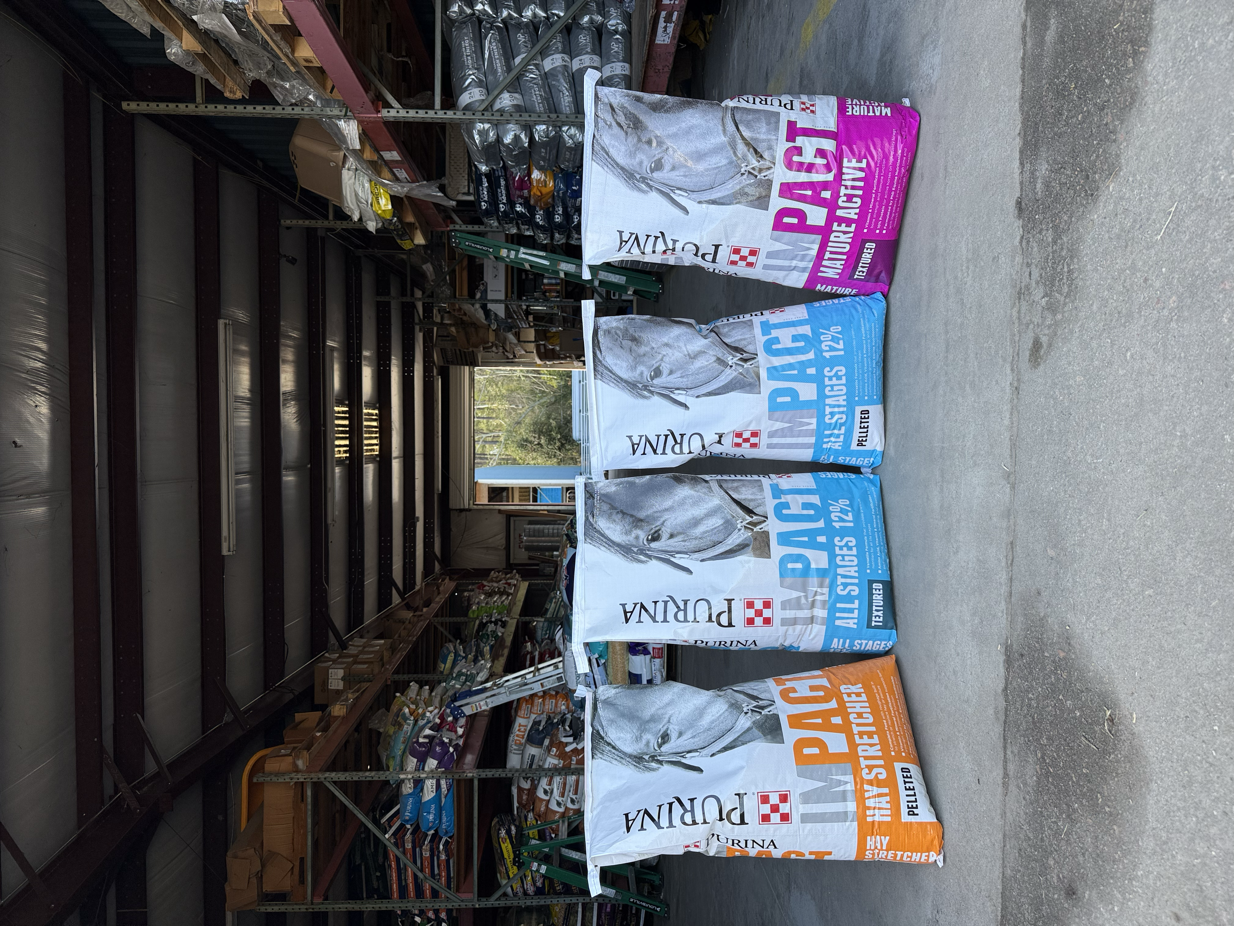 Several large bags of Purina horse feed labeled 'All Stages 12%' and 'Hay Stretcher' stacked outside a storage building.