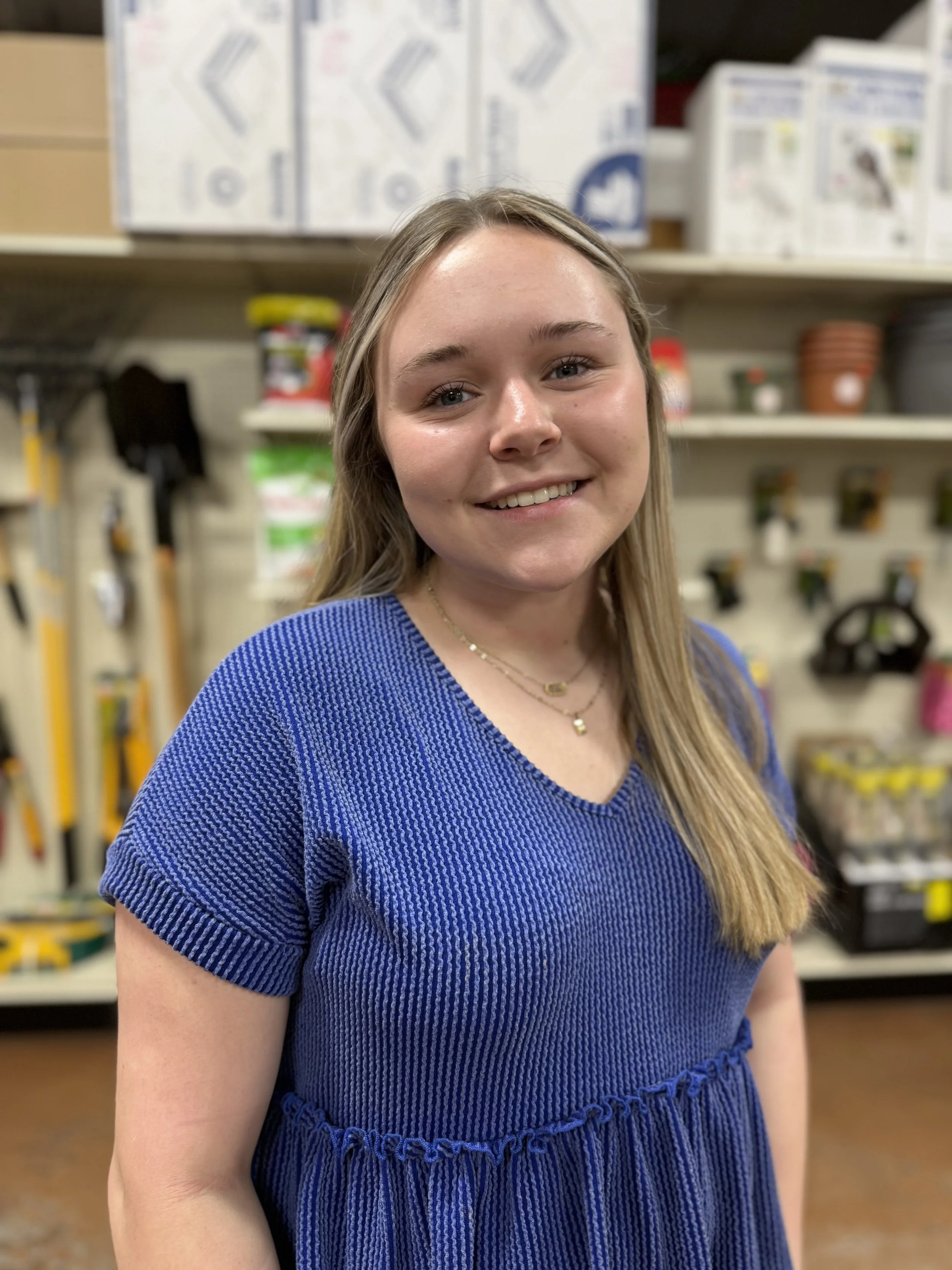 A young woman with long blonde hair smiling in an indoor hardware or garden store. She is wearing a blue knitted top and layered necklaces. Shelves with gardening supplies and tools are visible in the background.