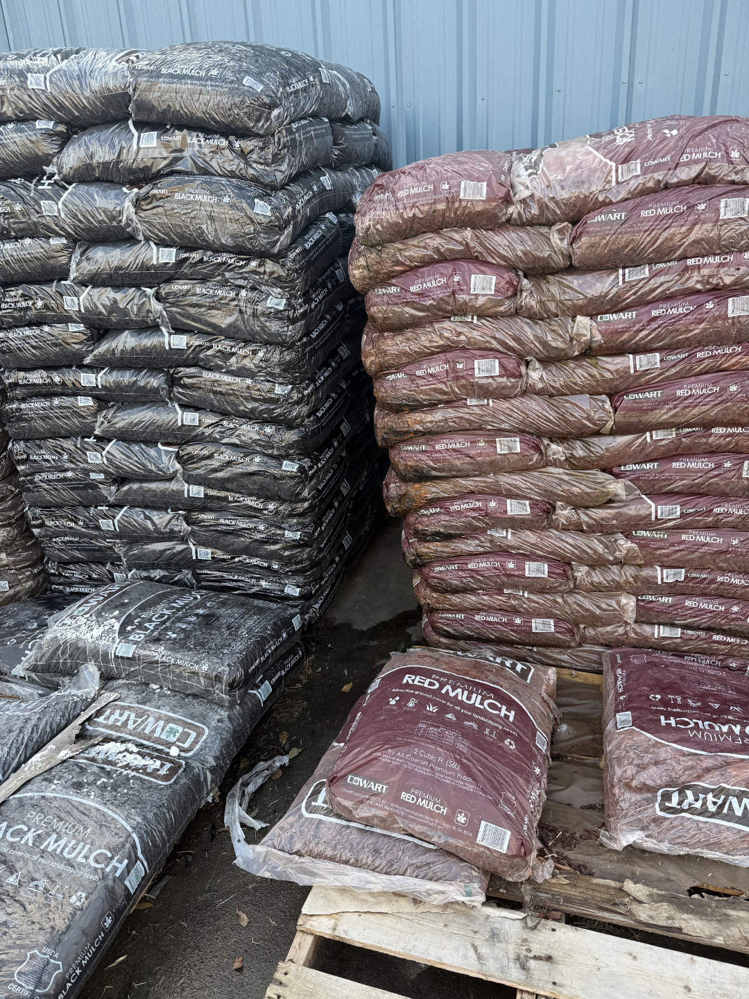 Stacks of black and red mulch bags stored outdoors against a blue metal wall, with some bags on a wooden pallet.