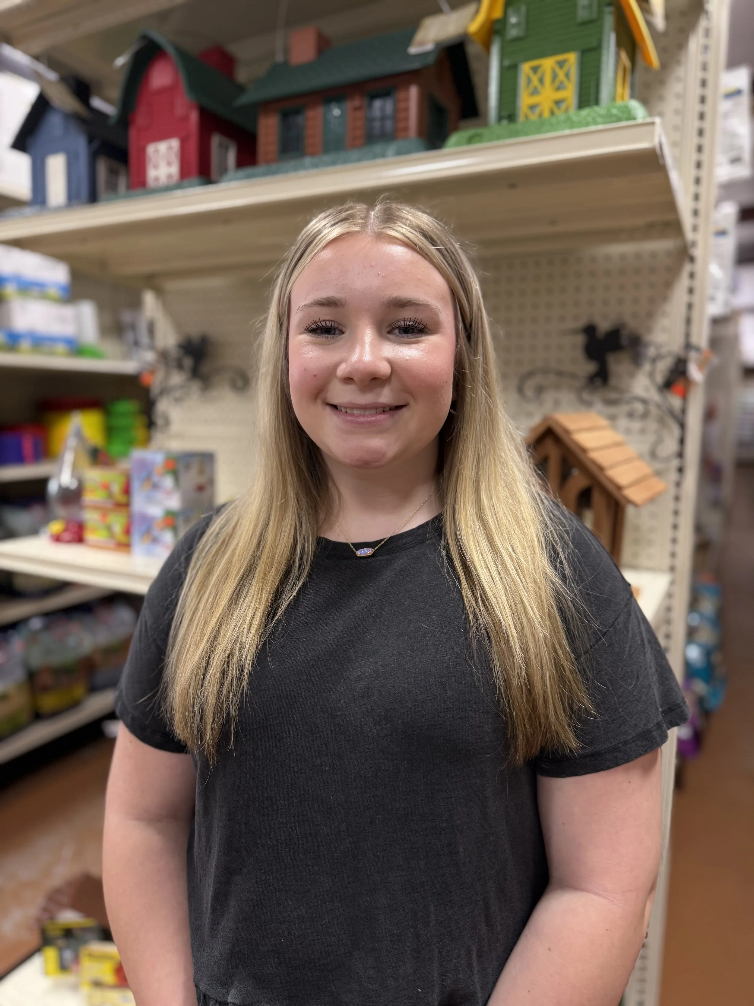 A young girl with long blonde hair smiling in a store aisle with toys and decorative items on shelves behind her.