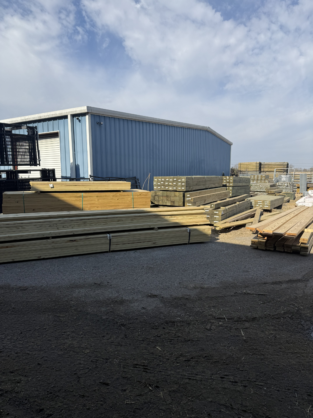 Outdoor storage yard with stacked lumber and wood planks in front of a blue metal building, under a cloudy sky.