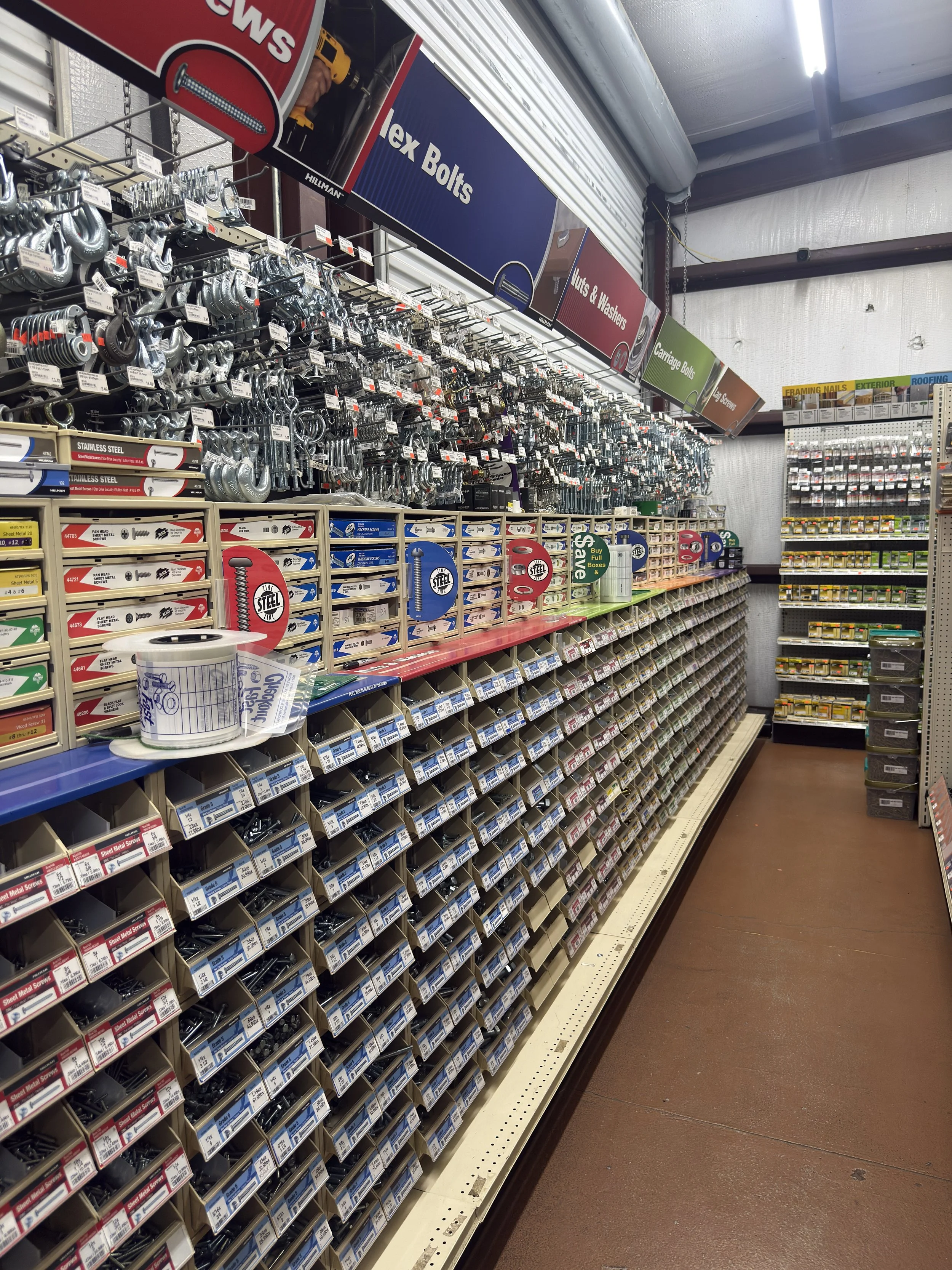 A hardware store aisle stocked with screws, bolts, nuts, and washers. Items are organized in small bins along the shelves, with signs indicating the types of fasteners, such as Tek Bolts, Nuts & Washers, and Carriage Bolts. There are additional supplies on a pegboard on the right.