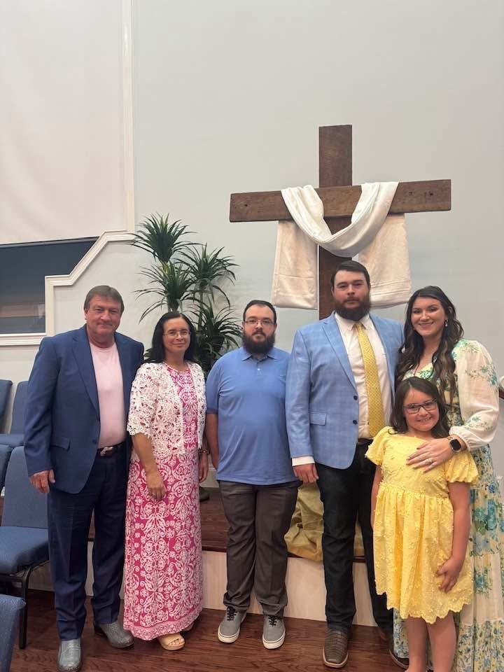 Group of six people, three men and three women, standing inside a church in front of a large wooden cross with a white cloth draped over it, during a religious ceremony or gathering.