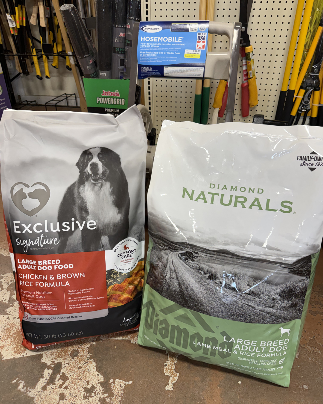 Two bags of large breed adult dog food, one labeled 'Exclusive Signature' chicken and brown rice formula, and the other labeled 'Diamond Naturals' lamb meal and rice formula, displayed on a store floor with gardening tools in the background.