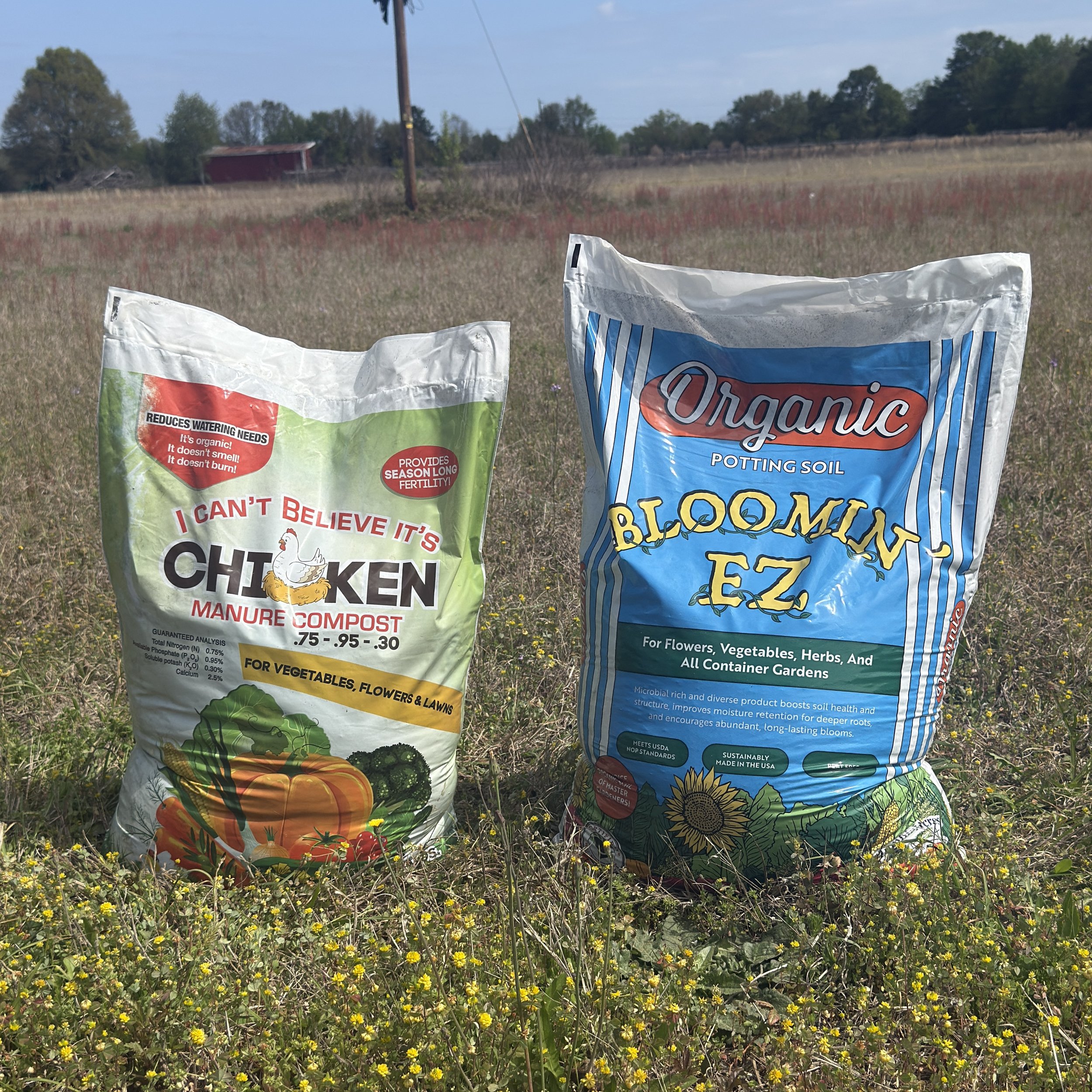 Two bags of gardening soil and compost placed in a field of small yellow flowers with trees visible in the background.