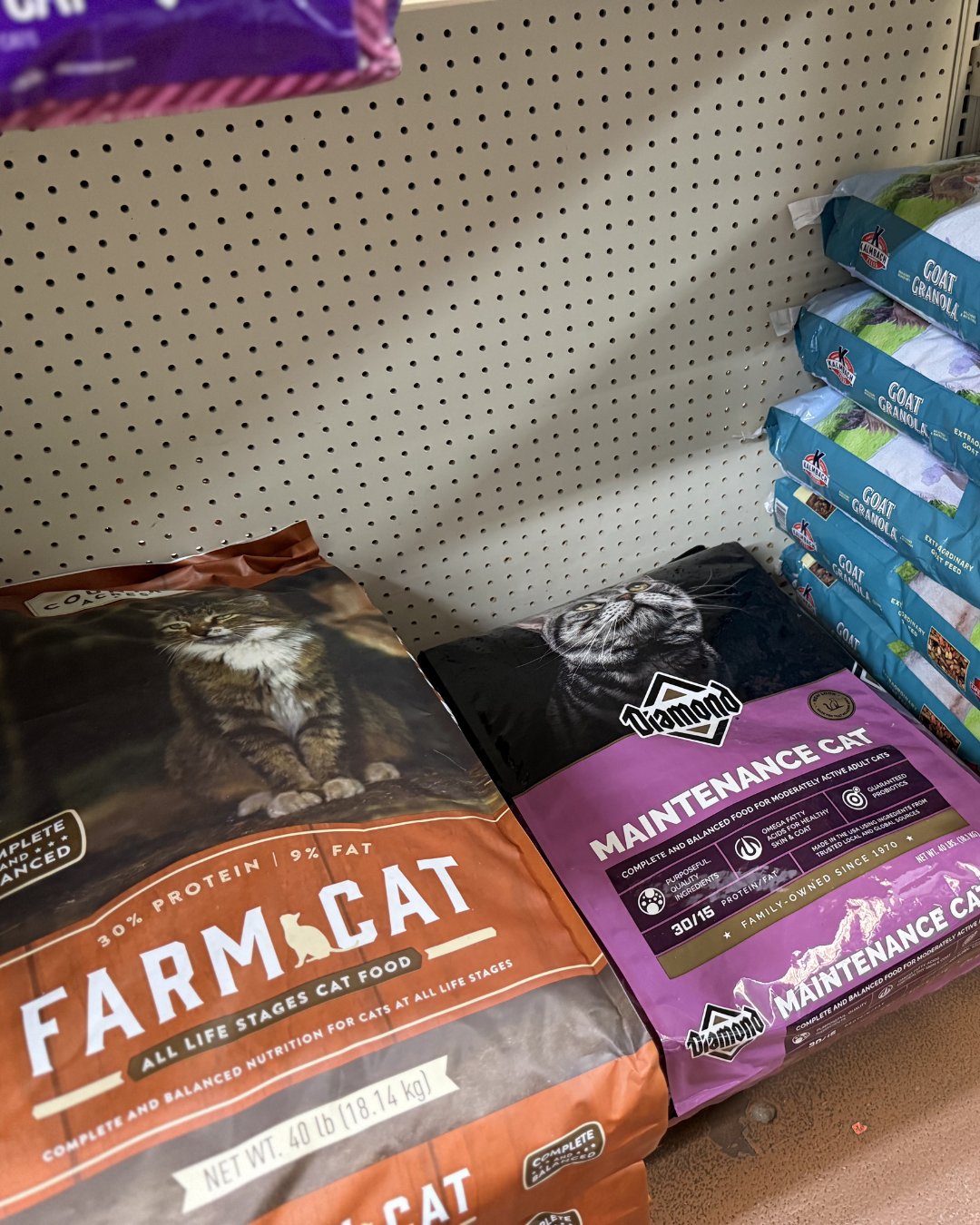 Cat food bags on a store shelf, including Farm Cat and Maintenance Cat brands, with a green bag of goat granola in the background.